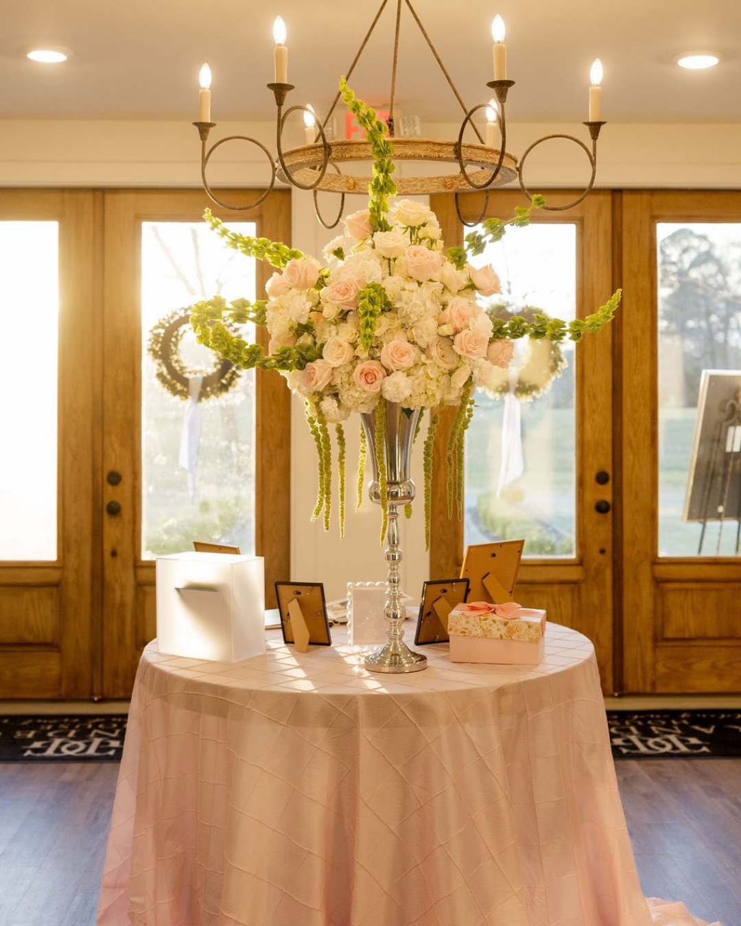 Elegant centerpiece with pink and white roses and green accents on a pink tablecloth, illuminated by a chandelier, in a well-lit room with large wooden framed windows.