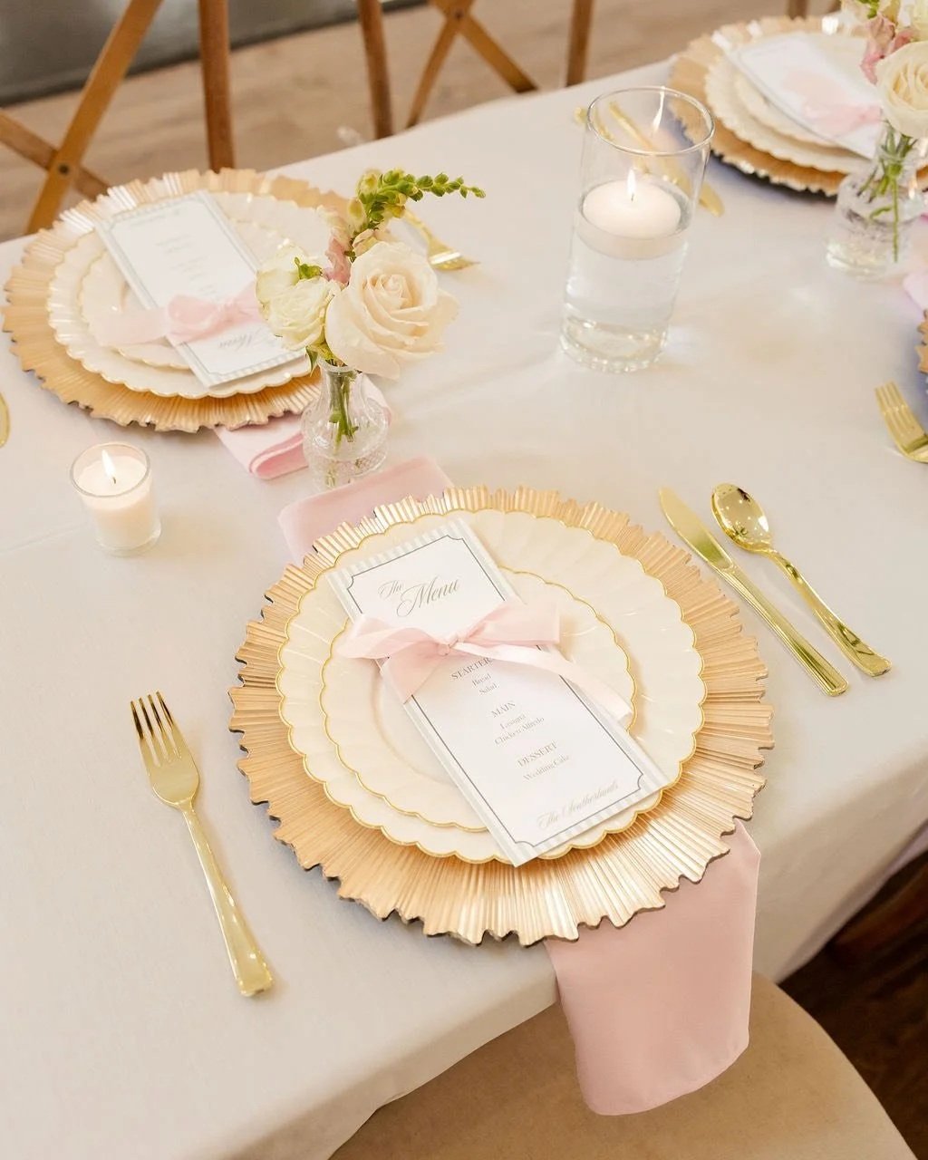 Elegant table setting with gold-rimmed plates, pink napkins tied with ribbons, gold utensils, candles, and a small floral centerpiece with white roses, set on a white tablecloth for a formal event.