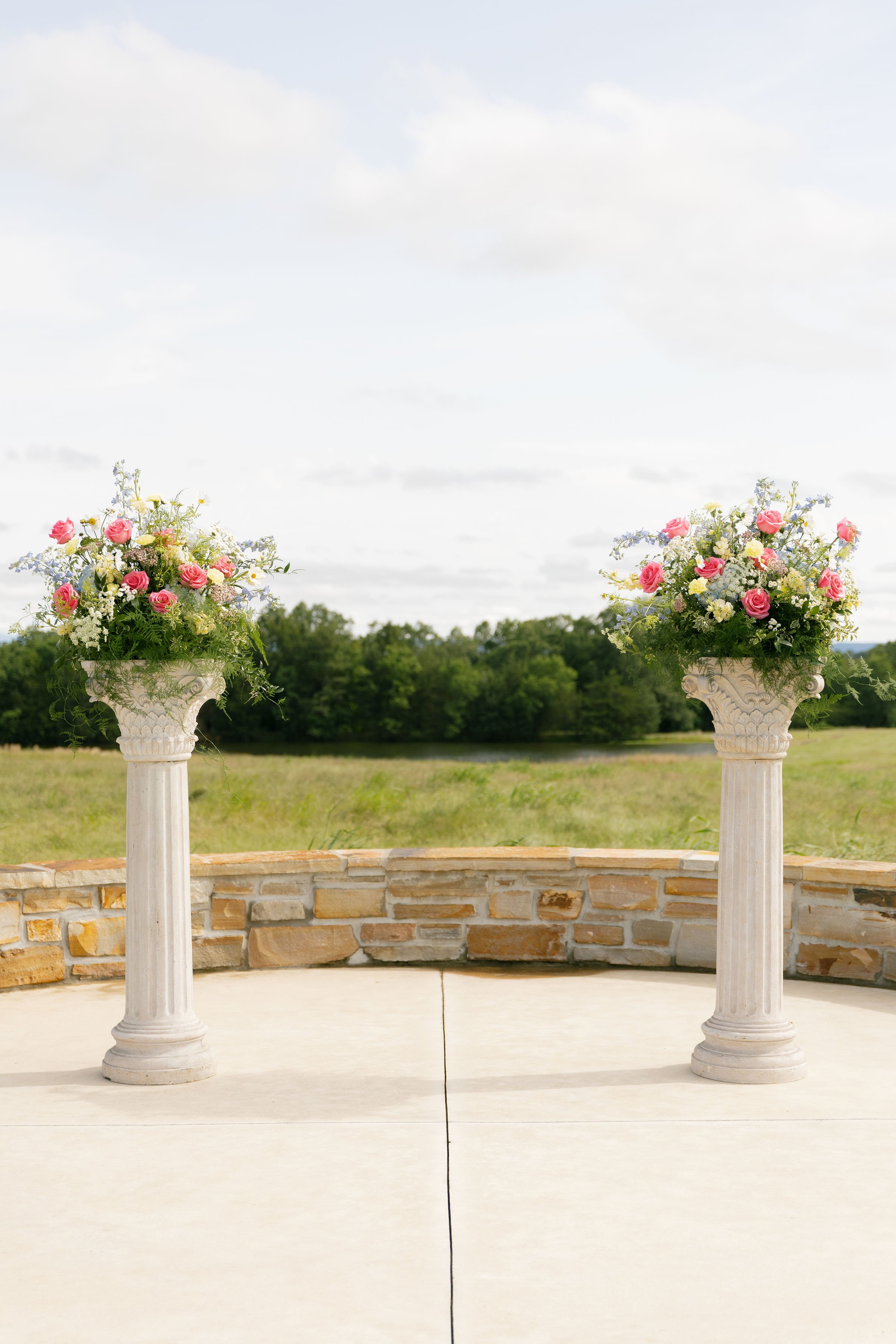 Two white stone pillars with floral arrangements featuring pink, yellow, and blue flowers, set against a background of green trees and cloudy sky.