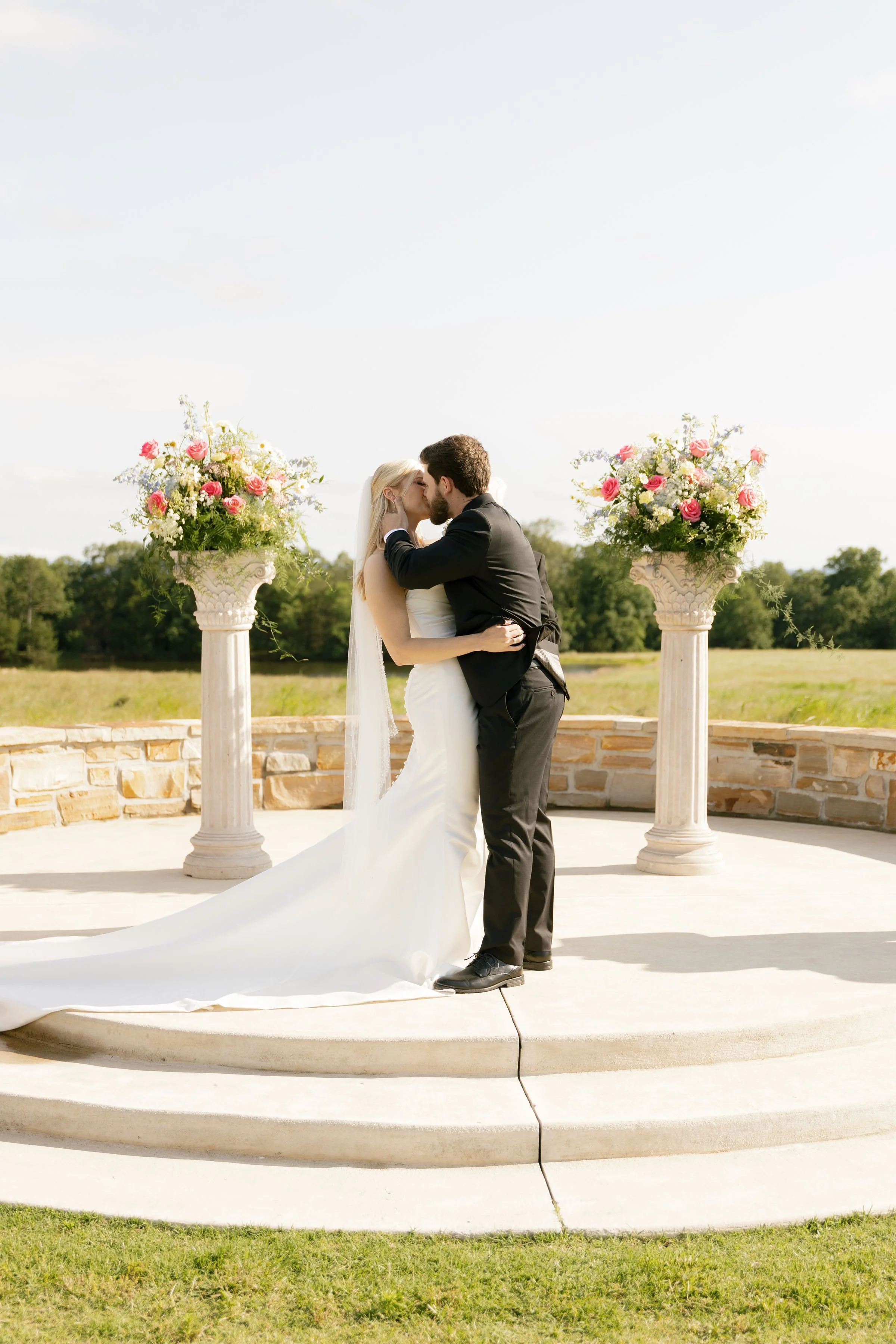 A newlywed couple sharing a kiss at an outdoor wedding ceremony, standing on a step in front of two large floral arrangements on tall white columns, with a green grassy field and trees in the background.