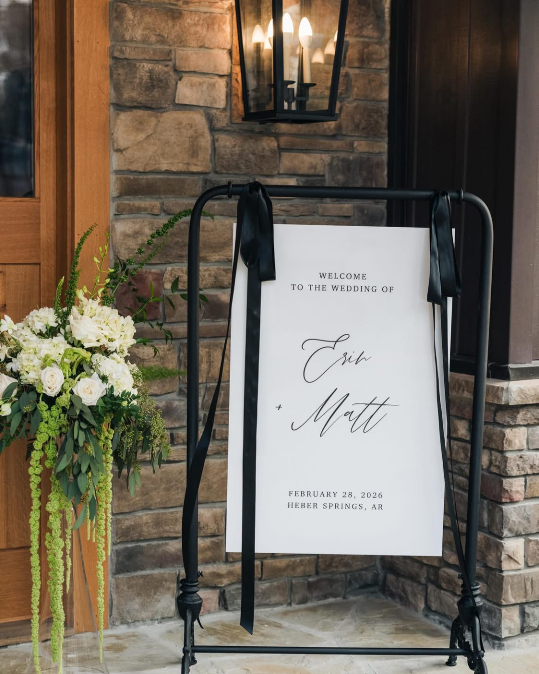 Wedding welcome sign with black ribbons, standing on a metal frame next to a floral arrangement of white roses and greenery, against a brick wall and lantern lighting.
