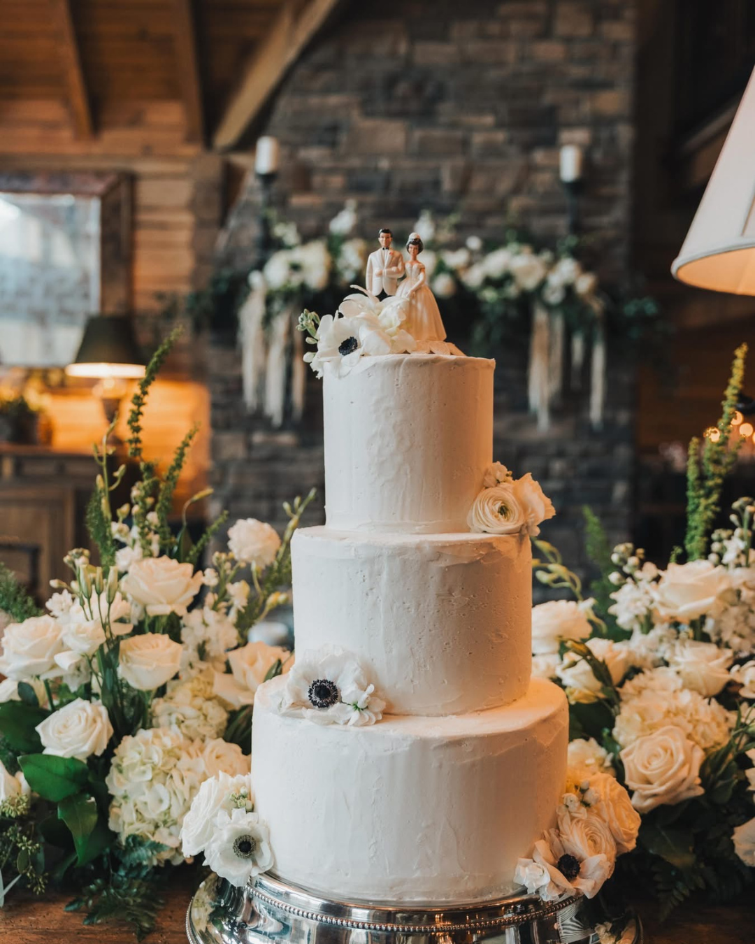 Three-tiered white wedding cake decorated with white flowers and a bride and groom topper, on a silver stand surrounded by white roses and greenery.