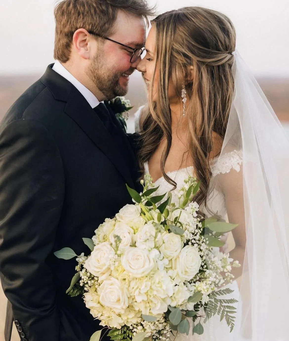 A bride and groom on their wedding day, touching foreheads and smiling, with the bride holding a large bouquet of white roses and greenery, and the bride wearing a lace dress with a veil and dangling earrings, and the groom in a black suit.