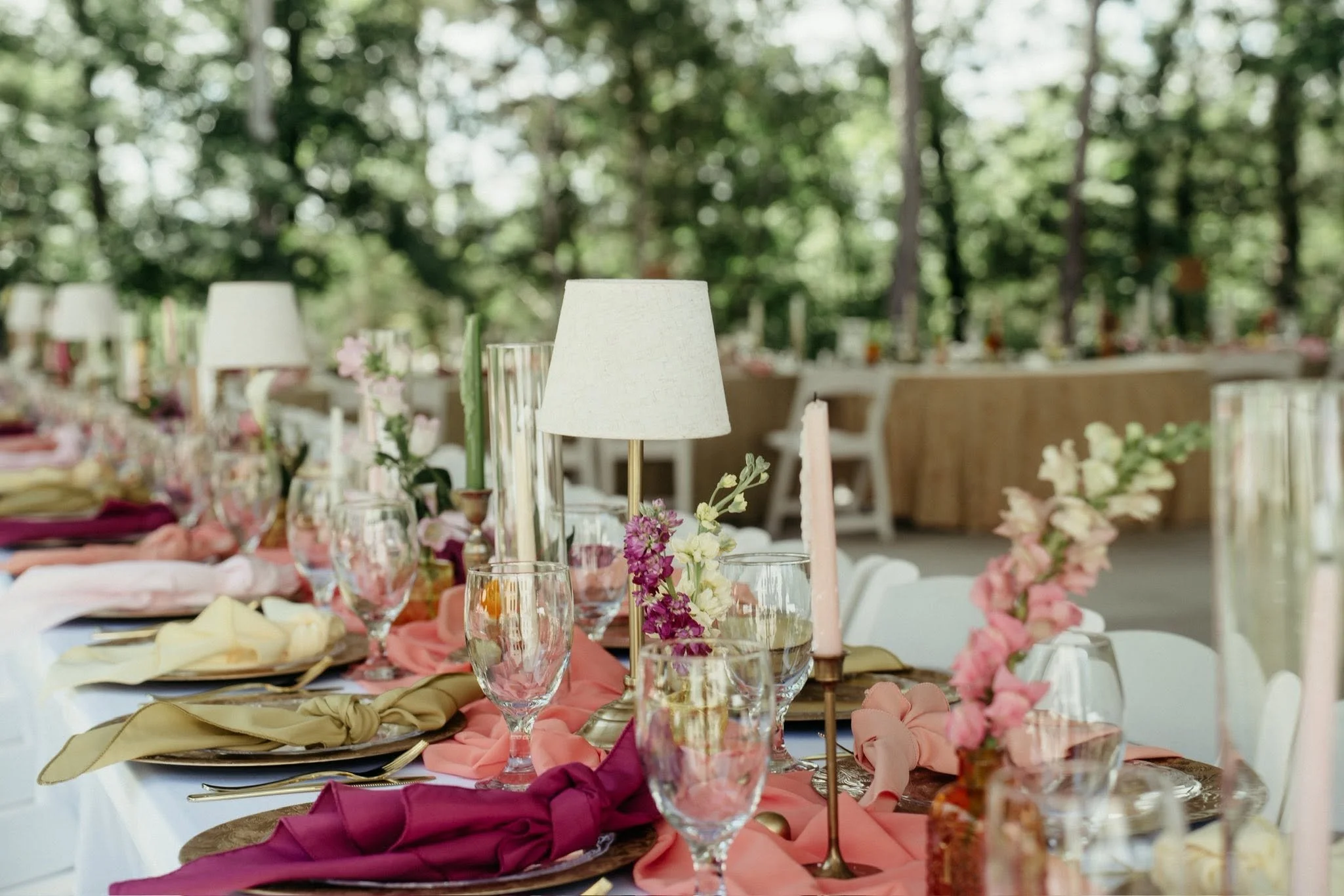 A decorated outdoor event table with floral arrangements, candle holders, glasses, and folded napkins, set in a natural green setting.