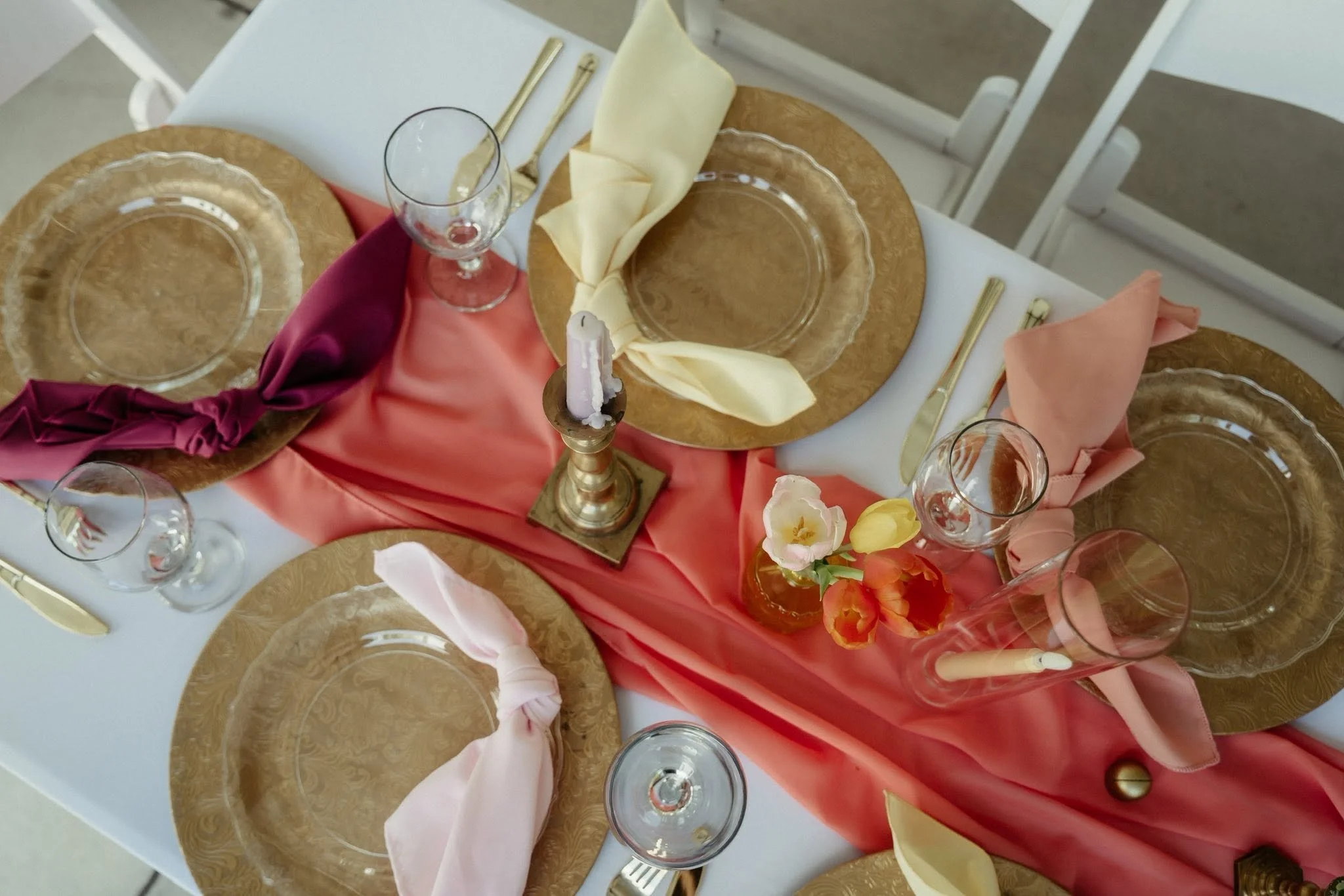 A dining table set with gold-rimmed glass plates, wine glasses, cutlery, and colorful napkins. A pink and coral fabric runner with floral accents decorates the table, which also features a brass candle holder with a single candle.