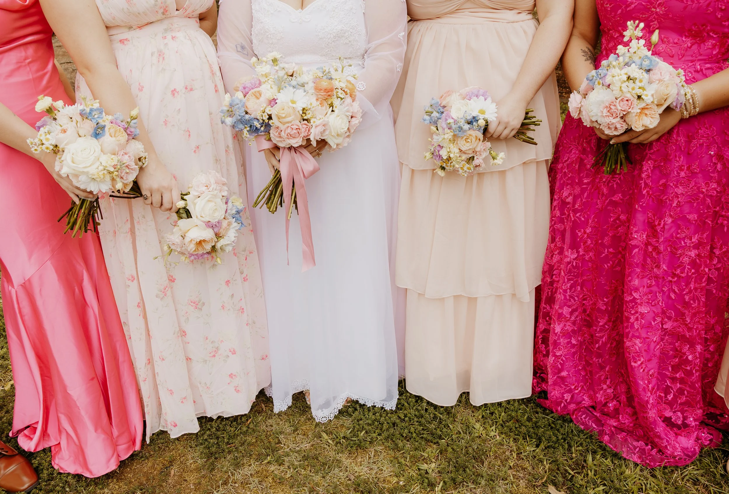 Close-up of women standing outdoors, wearing pastel-colored and floral dresses, holding bouquets of flowers with white, pink, blue, and purple blooms.