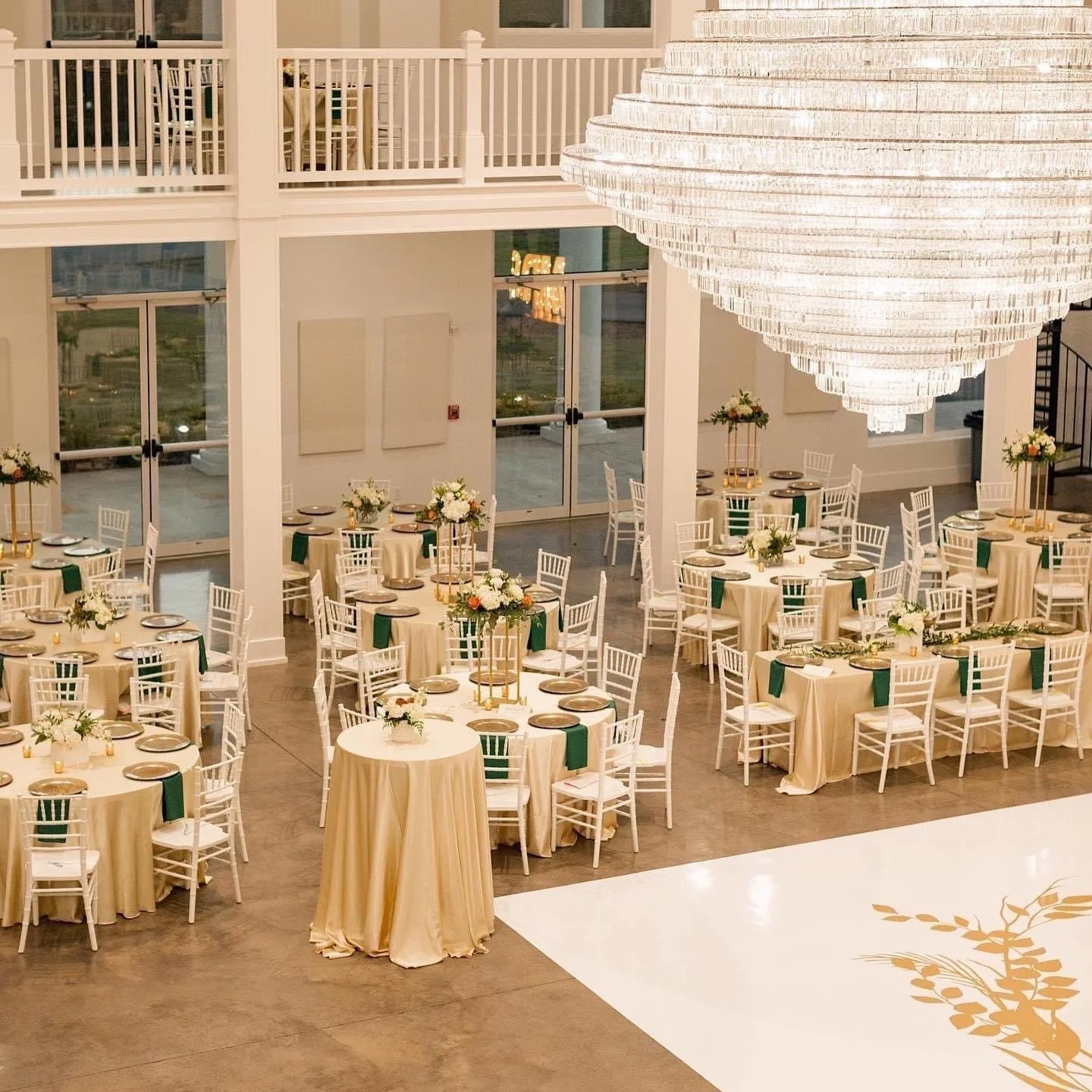 Elegant event venue with round tables decorated with floral centerpieces, set for a formal gathering with white chairs and gold accents, illuminated by a grand chandelier.