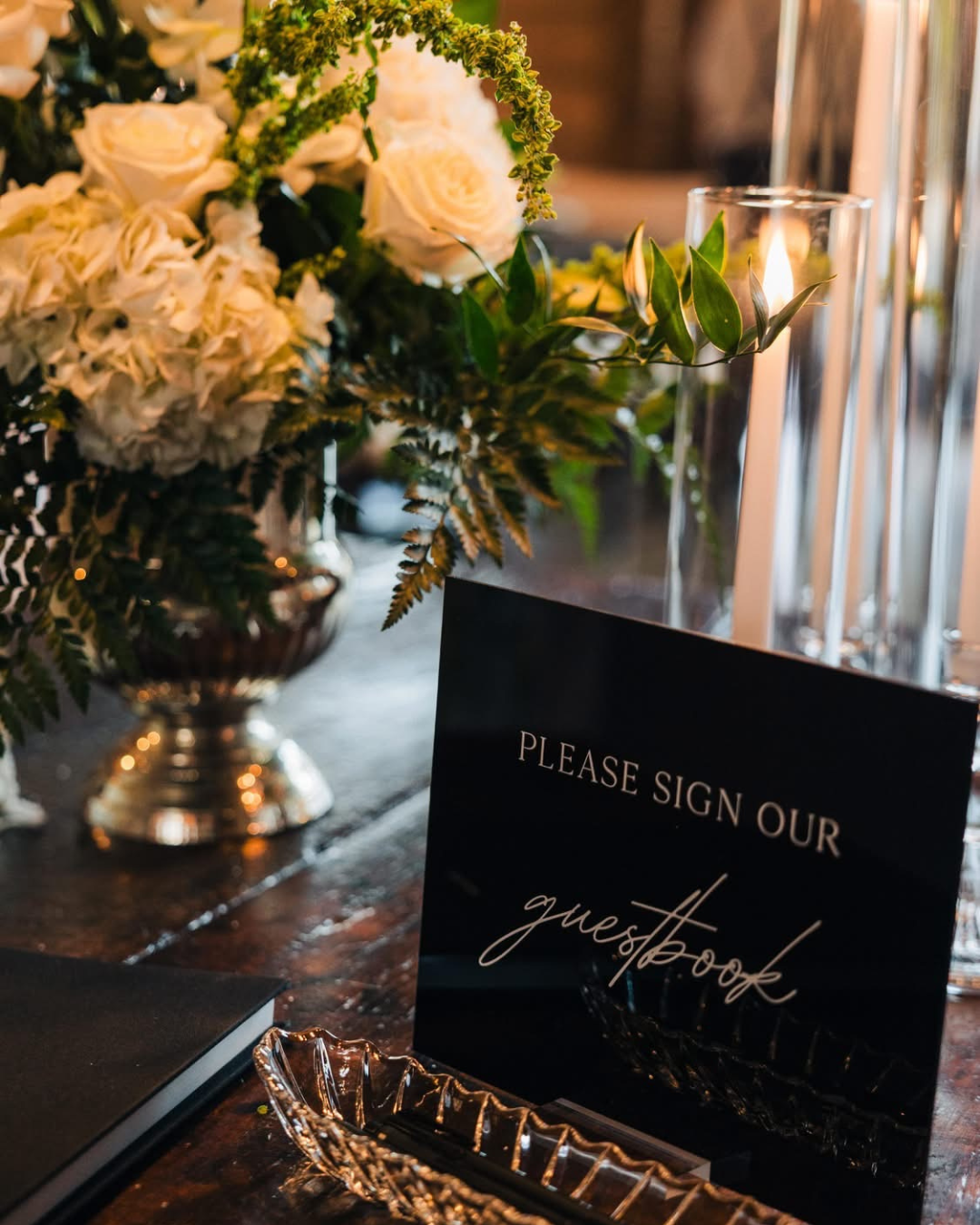 A floral arrangement with white roses and greenery in a silver vase, a black guestbook sign, and a lit candle in a glass holder on a dark wooden table.