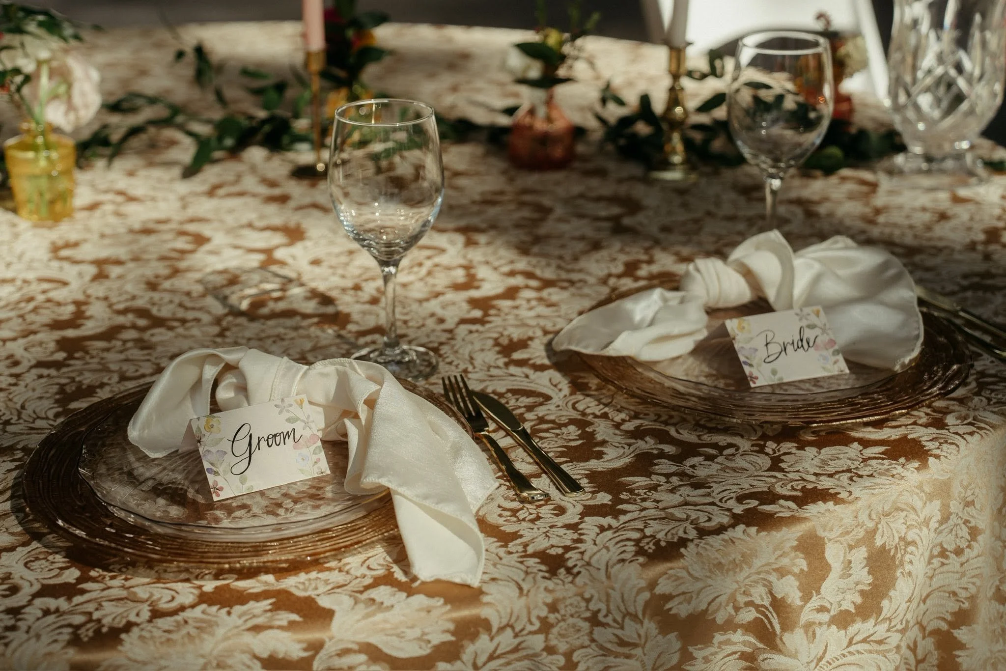 Wedding reception table setting with two glasses, decorative foliage, candlesticks, and place cards labeled 'Groom' and 'Bride' on a patterned tablecloth.