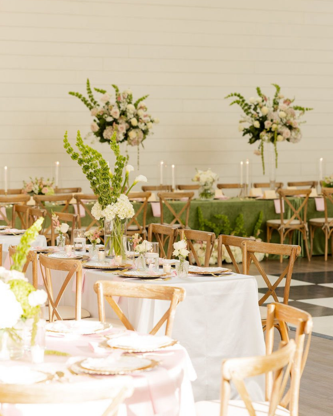 Elegant wedding reception setup with tables decorated with pink and white flowers, green foliage, and candles, with floral arrangements on a checkered floor and white wall background.
