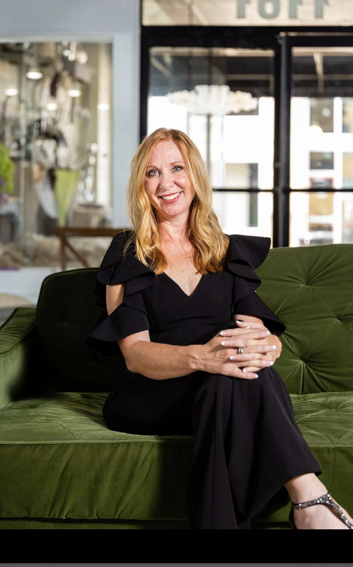A smiling woman with red hair and blue eyes sitting on a green velvet sofa inside a brightly lit café or restaurant, wearing a black dress with ruffled shoulders and silver shoes, with a blurred background of large windows and modern decor.