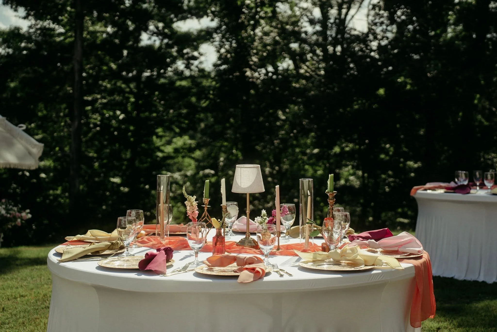 A round banquet table set for an outdoor event with pink and peach napkins, wine glasses, candleholders, and floral decorations, with lush green trees in the background.