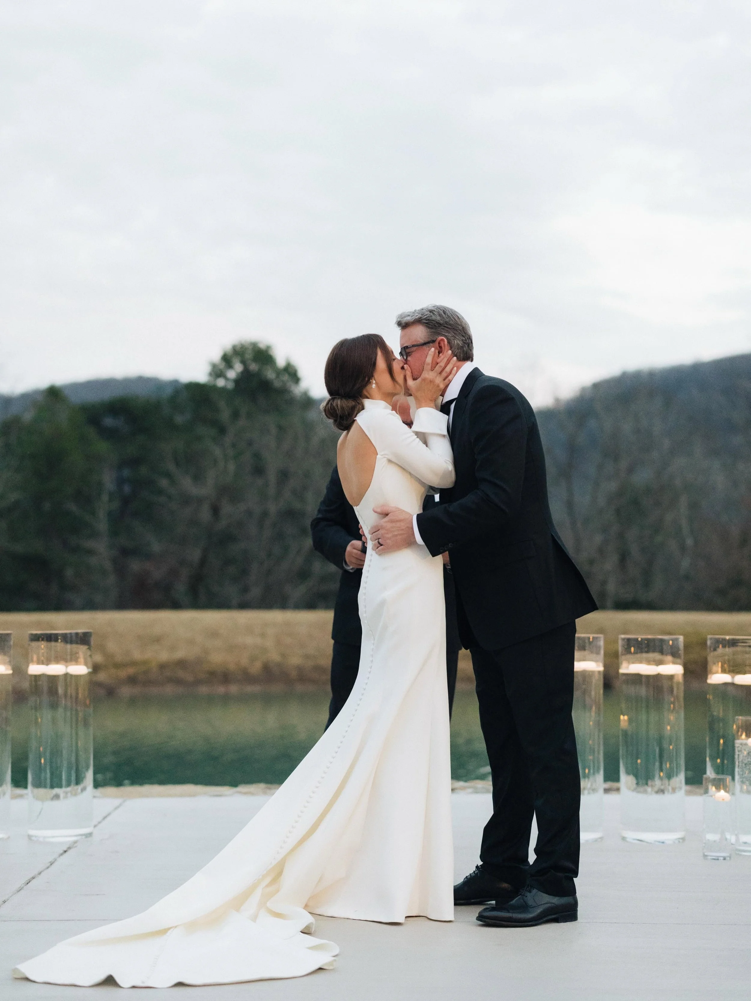 A couple sharing a kiss at their wedding ceremony outdoors, with a scenic nature background and water nearby, during what appears to be late afternoon or early evening.