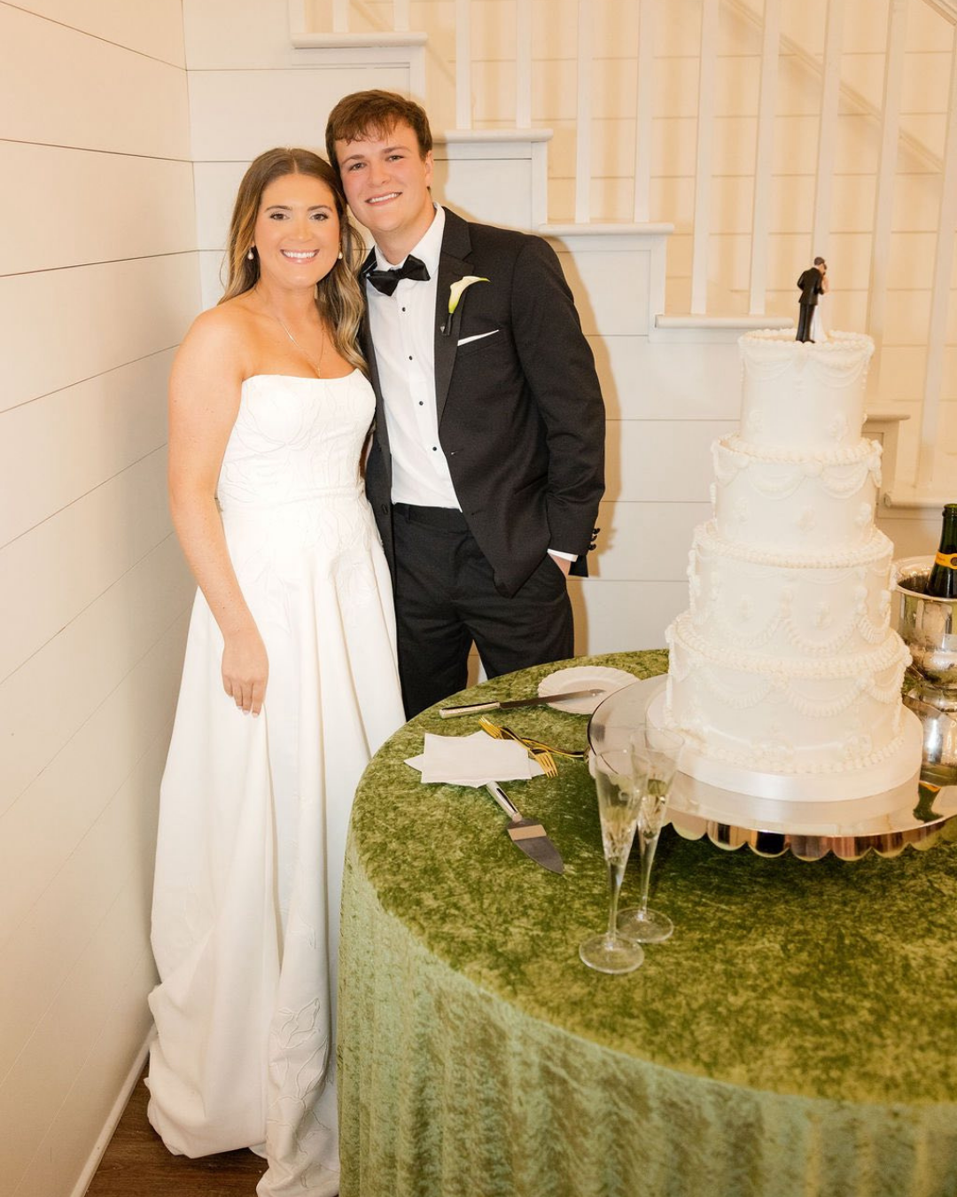 A bride and groom in wedding attire standing next to a wedding cake on a green table with champagne glasses and silverware.