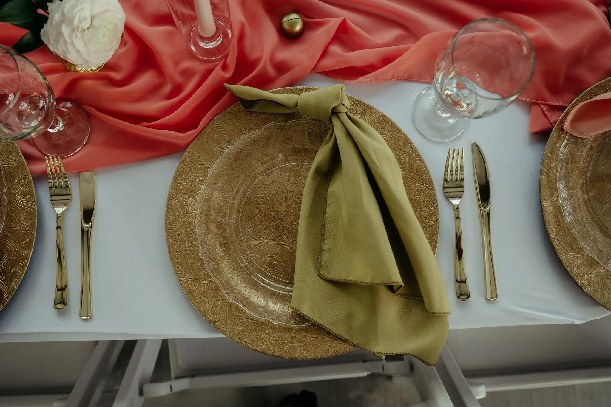 A table set for a formal event with gold-patterned plates, gold cutlery, clear glassware, a beige napkin folded at each place, and a coral-colored table runner with a white flower centerpiece and a small decorative orb.