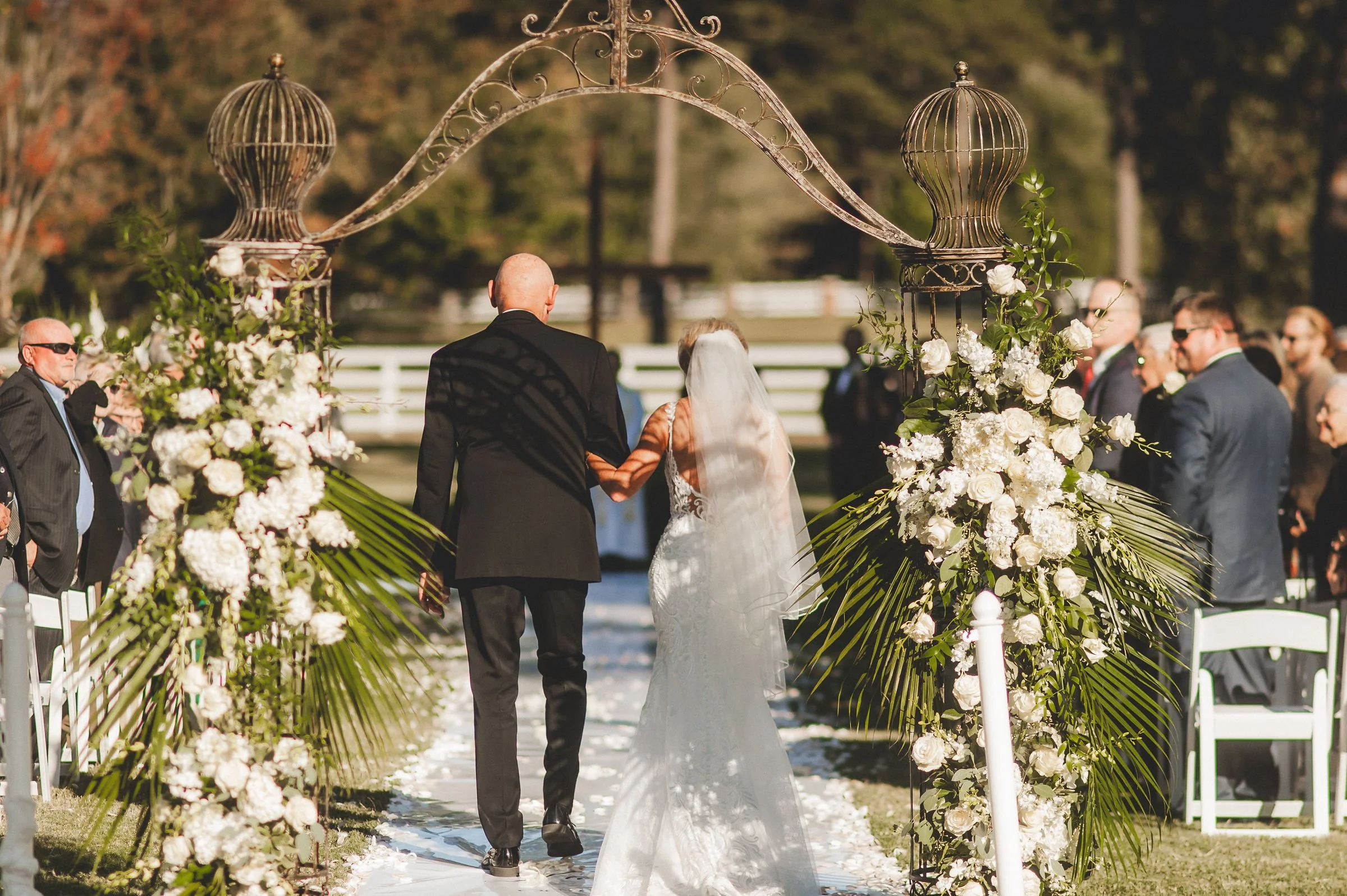A wedding ceremony outdoors with a bride and groom walking down the aisle surrounded by guests and floral decorations.