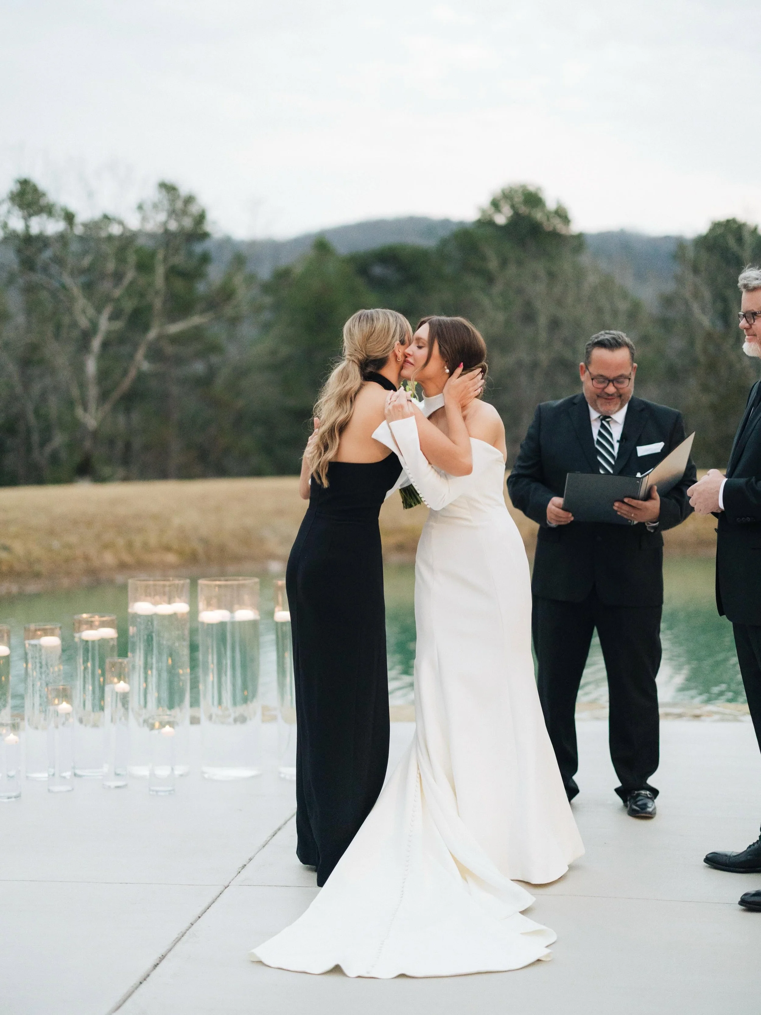 Two women, one in a white wedding dress and the other in a black dress, kiss during a wedding ceremony outdoors. Two men, dressed in suits, are standing nearby, with one reading from a folder. Candles are arranged on the ground nearby, and trees and 