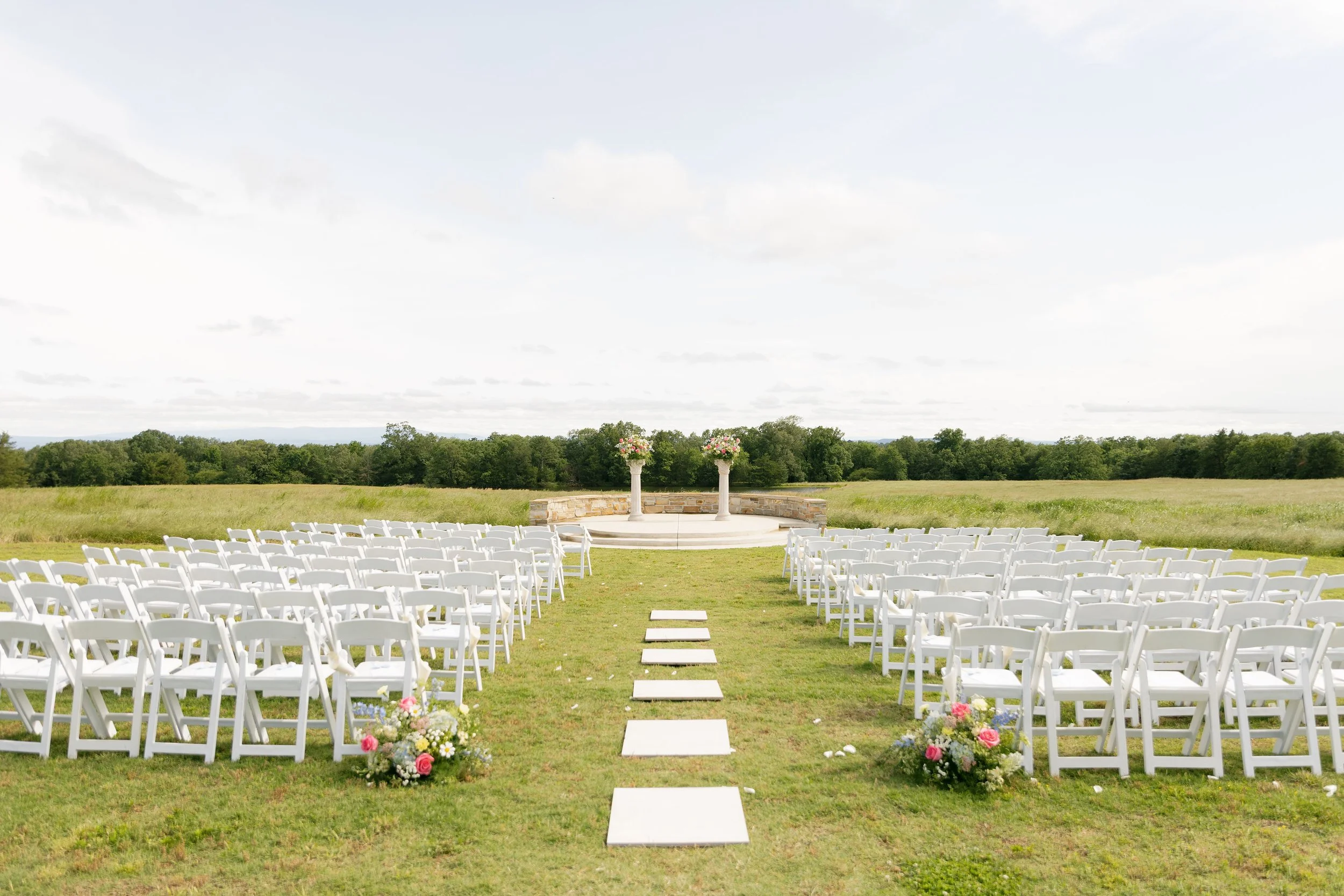 Outdoor wedding ceremony setup on a grassy field with white chairs arranged in rows, a stone altar with flower arrangements, and a scenic background of trees and cloudy sky.