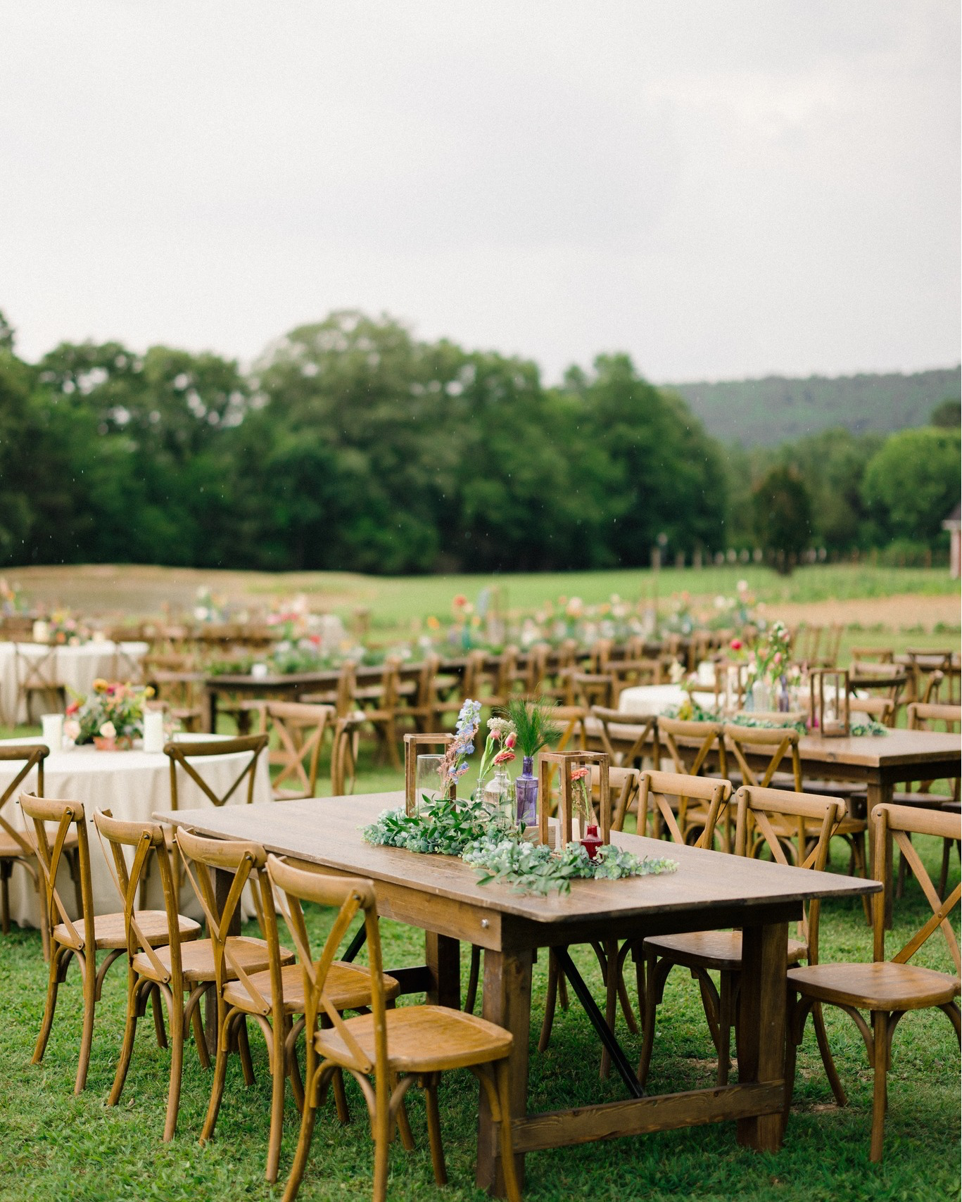 Outdoor wedding reception setup with wooden tables and chairs, decorated with floral centerpieces and candles, on a grassy field with trees and overcast sky in the background.