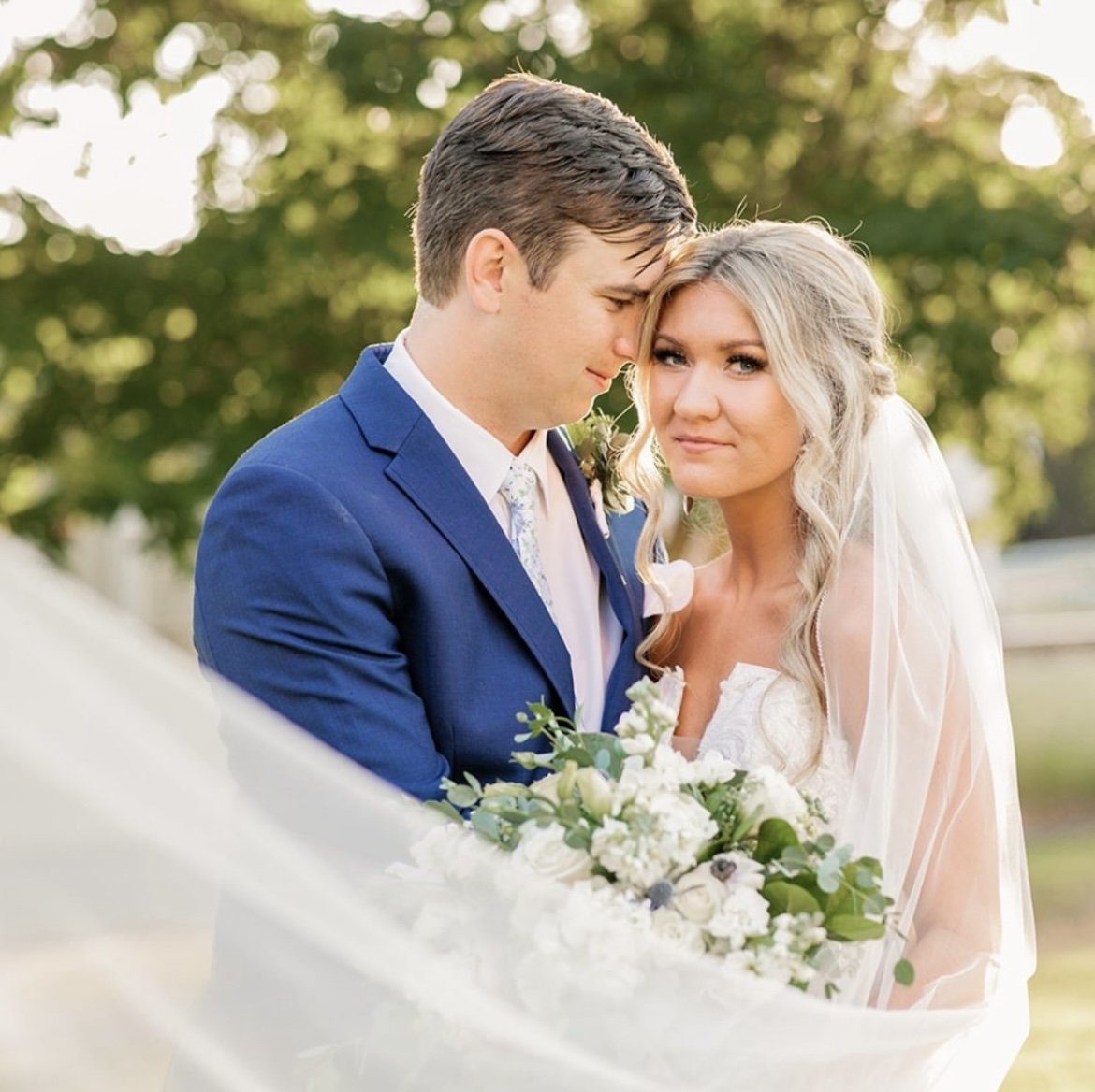 A bride and groom on their wedding day outdoors, with the groom in a blue suit and the bride holding a bouquet of white flowers and greenery, standing close together in front of green trees.