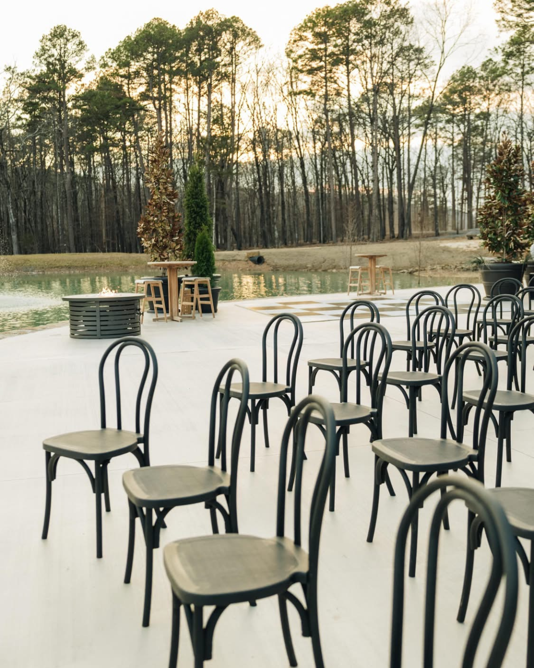 Outdoor event space with black chairs arranged on a light-colored floor, a pond surrounded by trees in the background, and small round tables overlooking the water during sunset.