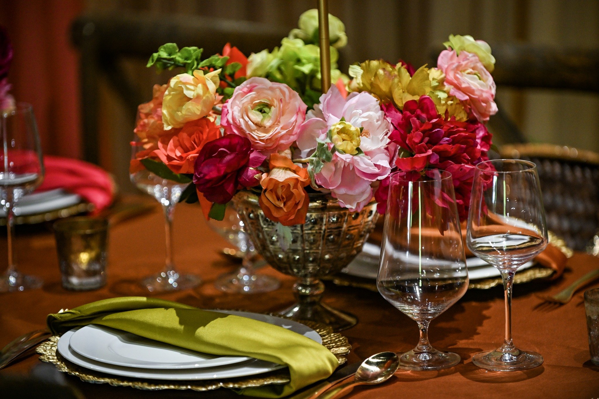 A centerpiece of colorful roses in a glass bowl on a dining table, surrounded by wine glasses, plates with a green napkin, and a gold-colored placemat.