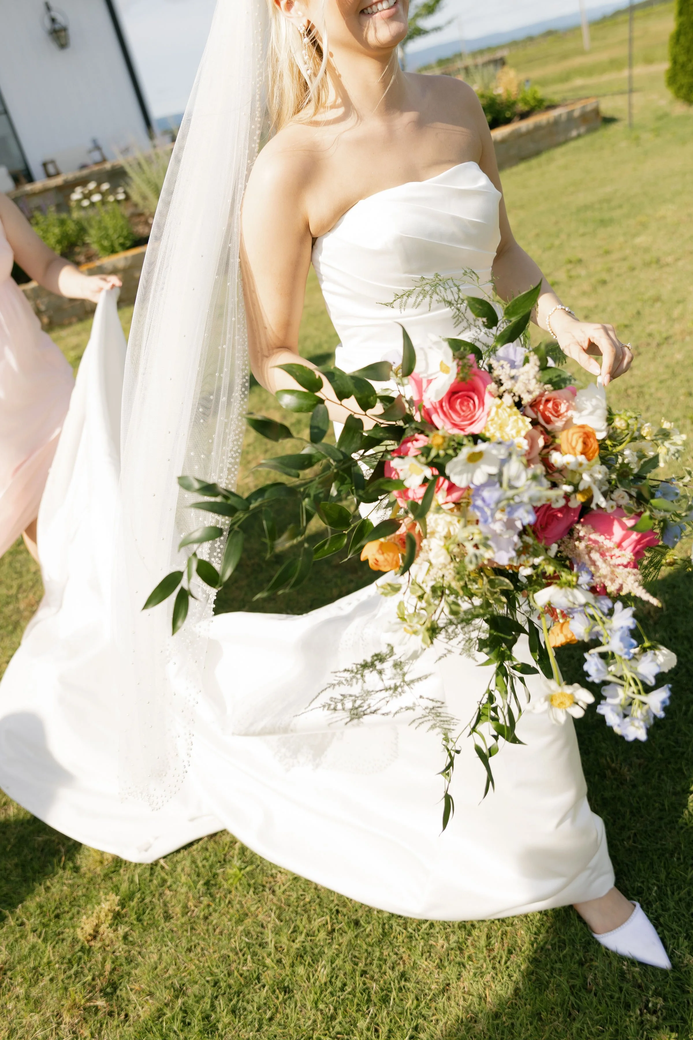 A woman in a strapless white wedding dress holding a vibrant bouquet of pink, orange, white, and purple flowers with green foliage, standing on grass outdoors, smiling.