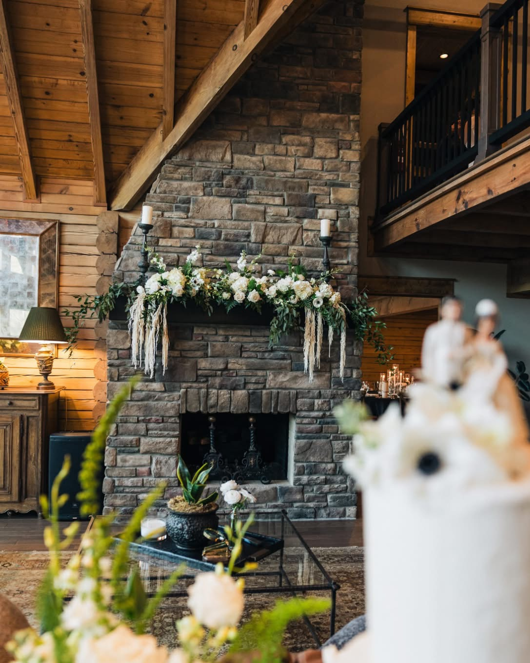 A rustic living room with a large stone fireplace decorated with white flowers and greenery, two tall candles on the mantle, and a small table with a potted plant in front of it. Part of a wedding or celebration setup is visible on the right side.