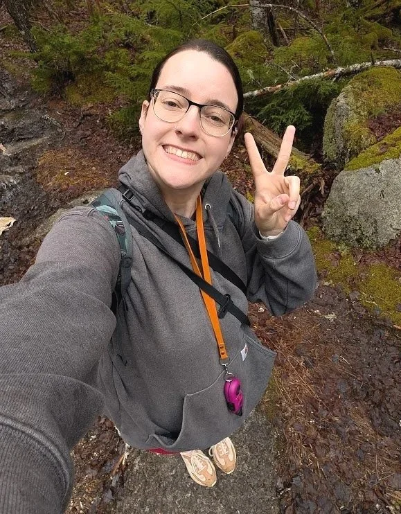 A young woman with glasses smiling and making a peace sign while hiking in a forest.
