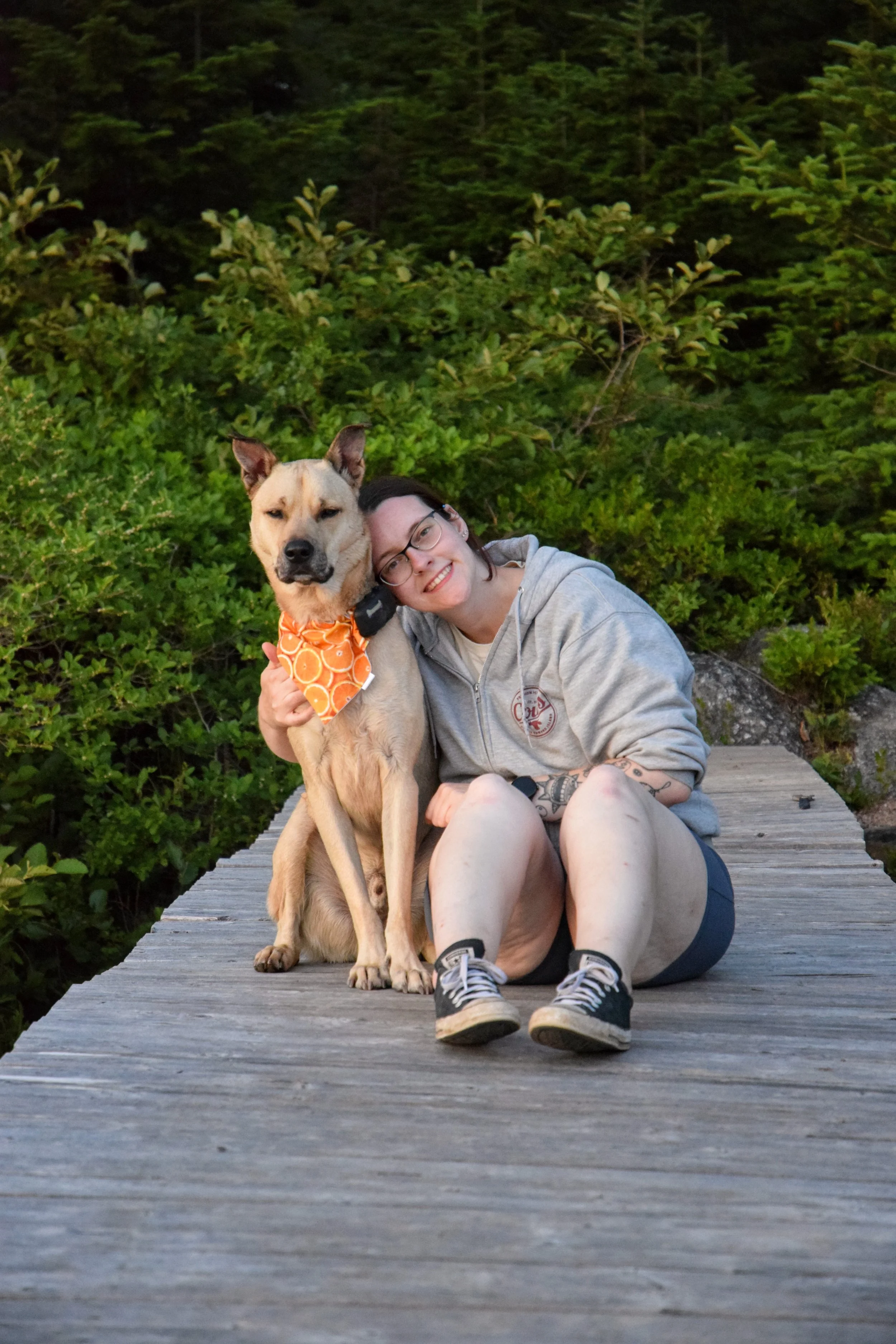 A person with glasses and a gray hoodie sitting on a wooden dock next to a large dog wearing an orange bandana, with lush green foliage in the background.