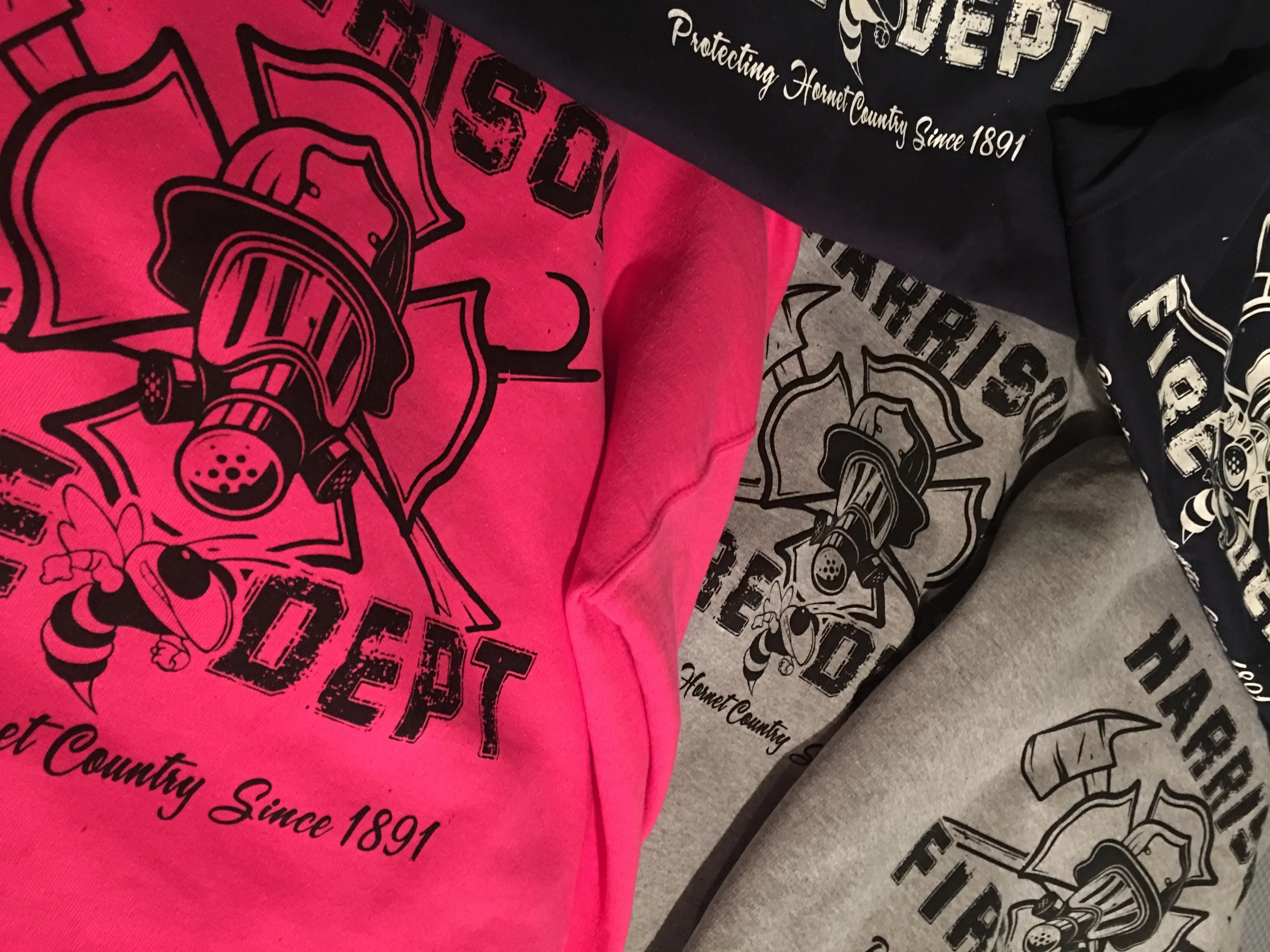 Close-up of multiple fabric shirts with a logo of a helmet, a helmet with a mouthpiece, and the text, 'Protecting Hometowne Since 1891', in black, white, and pink colors.