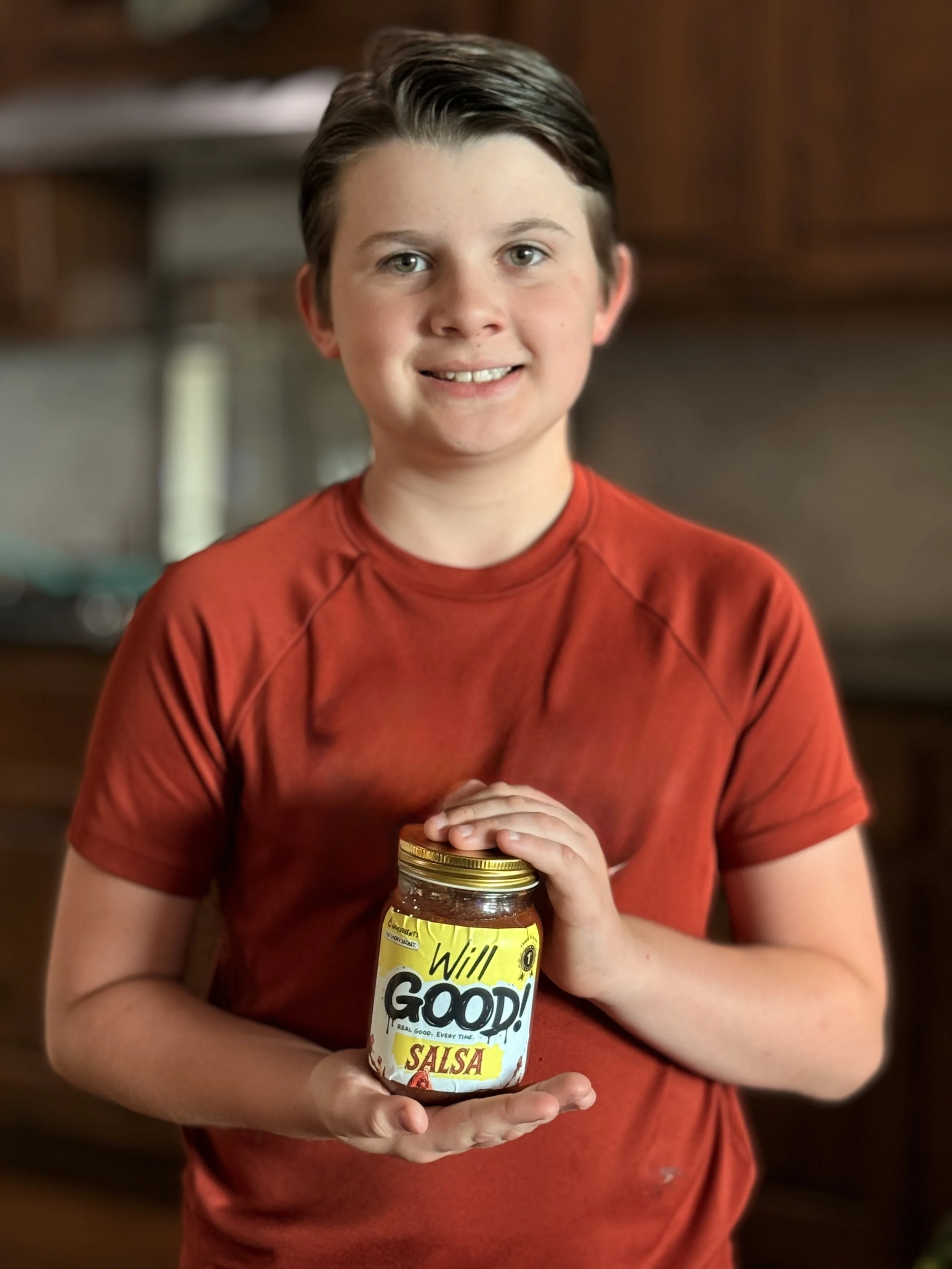 A young boy with short brown hair, wearing a red shirt, holding a jar of salsa with both hands, standing indoors with blurred wooden cabinets and counter in the background.