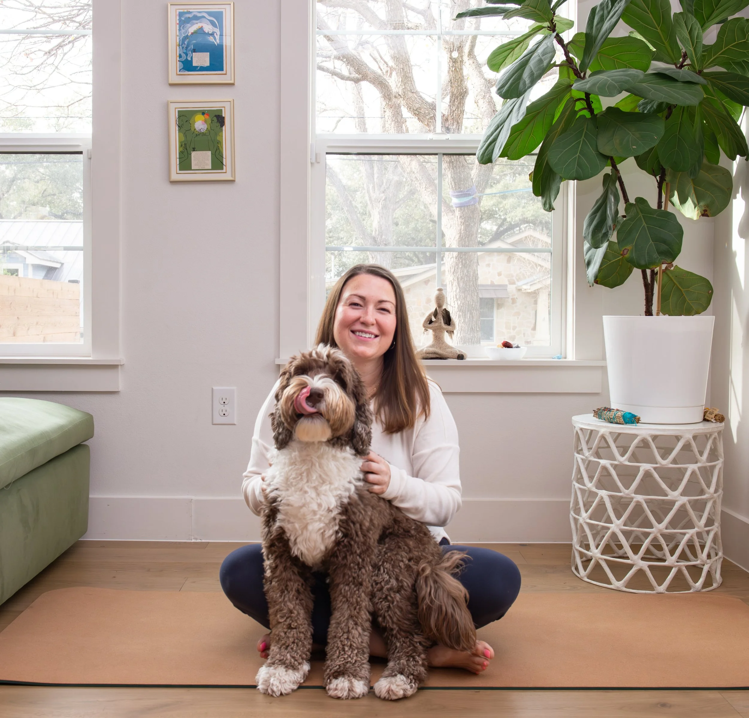 A woman with long brown hair smiling while holding a large fluffy dog with brown and white fur, seated in a cross-legged pose on a cushion in front of a window with a large leafy plant on her right. The background features framed artwork on the white wall and a view of trees outside.