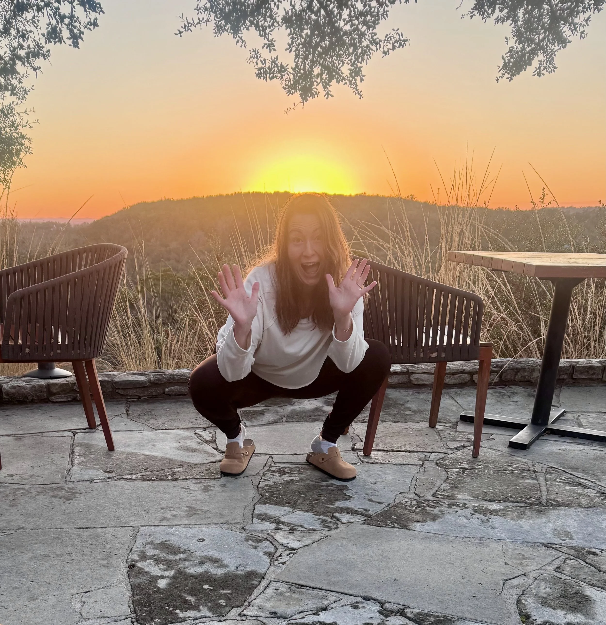 A woman squatting on a concrete patio at sunset, smiling and waving with both hands. There are outdoor patio chairs and a small table around her, with hills and a sunset sky in the background.