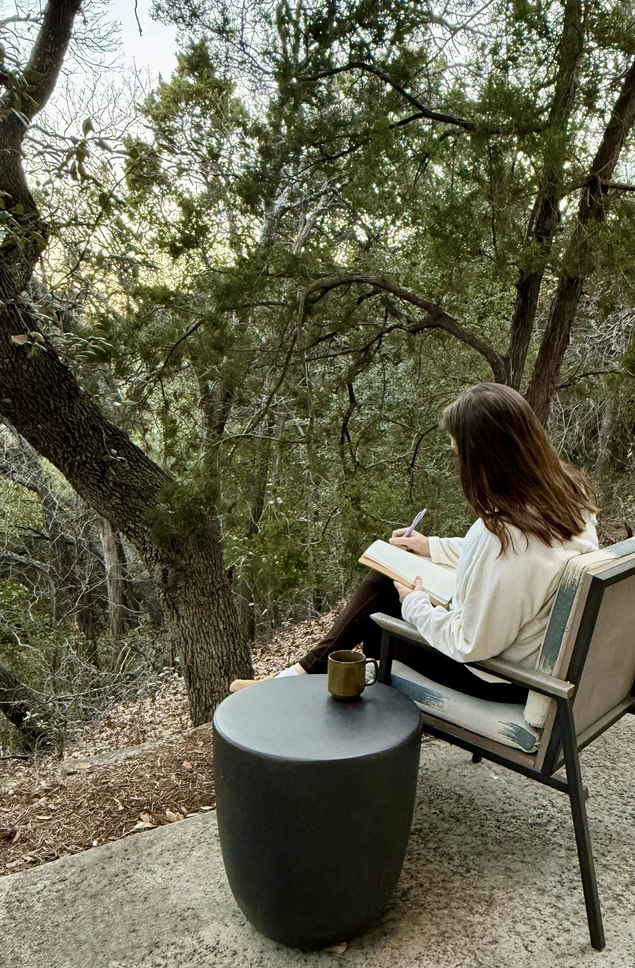 Person with brown hair wearing a white hoodie sitting on a wooden chair, writing in a notebook outdoors, with trees and foliage surrounding the area.