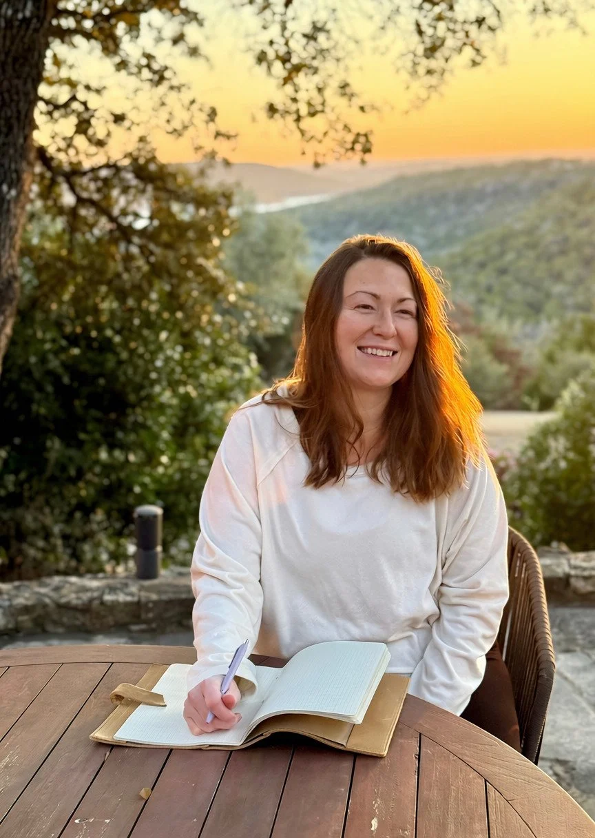 A woman with long brown hair smiling at a table outdoors during sunset, with a notebook and pen in front of her, surrounded by green trees and a scenic landscape.