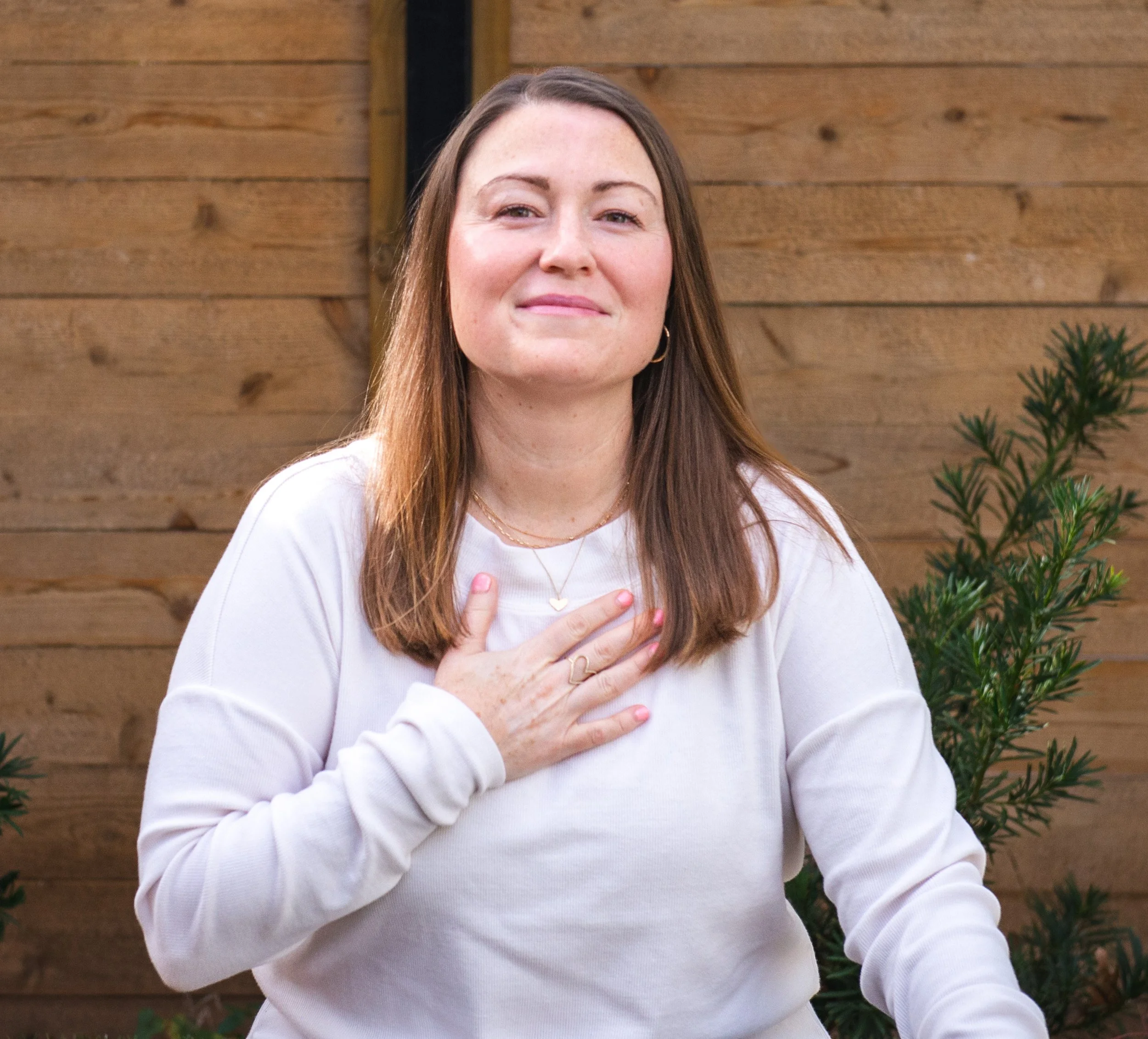 A woman with long brown hair wearing a light-colored sweater, standing outdoors against a wooden fence, with her right hand placed over her chest.