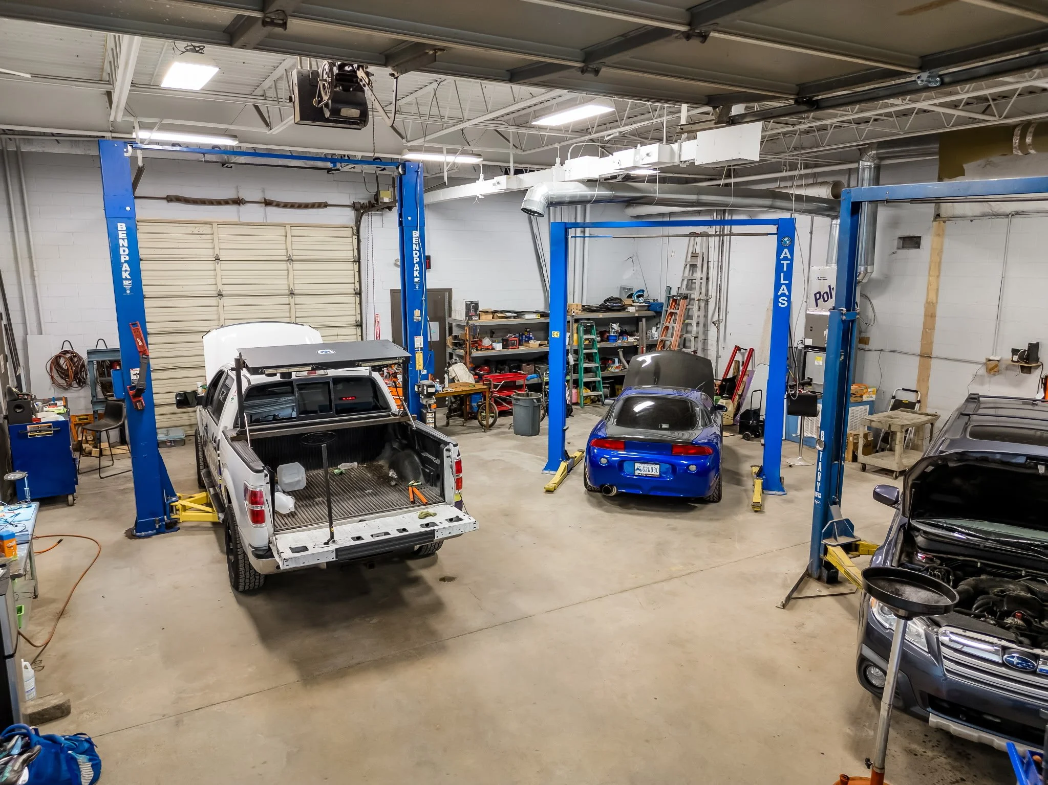 Auto repair shop with two car lifts; one lift has a partially assembled white pickup truck, the other has a blue sports car. Shelves with tools and equipment, several cars, and garage tools in the background.
