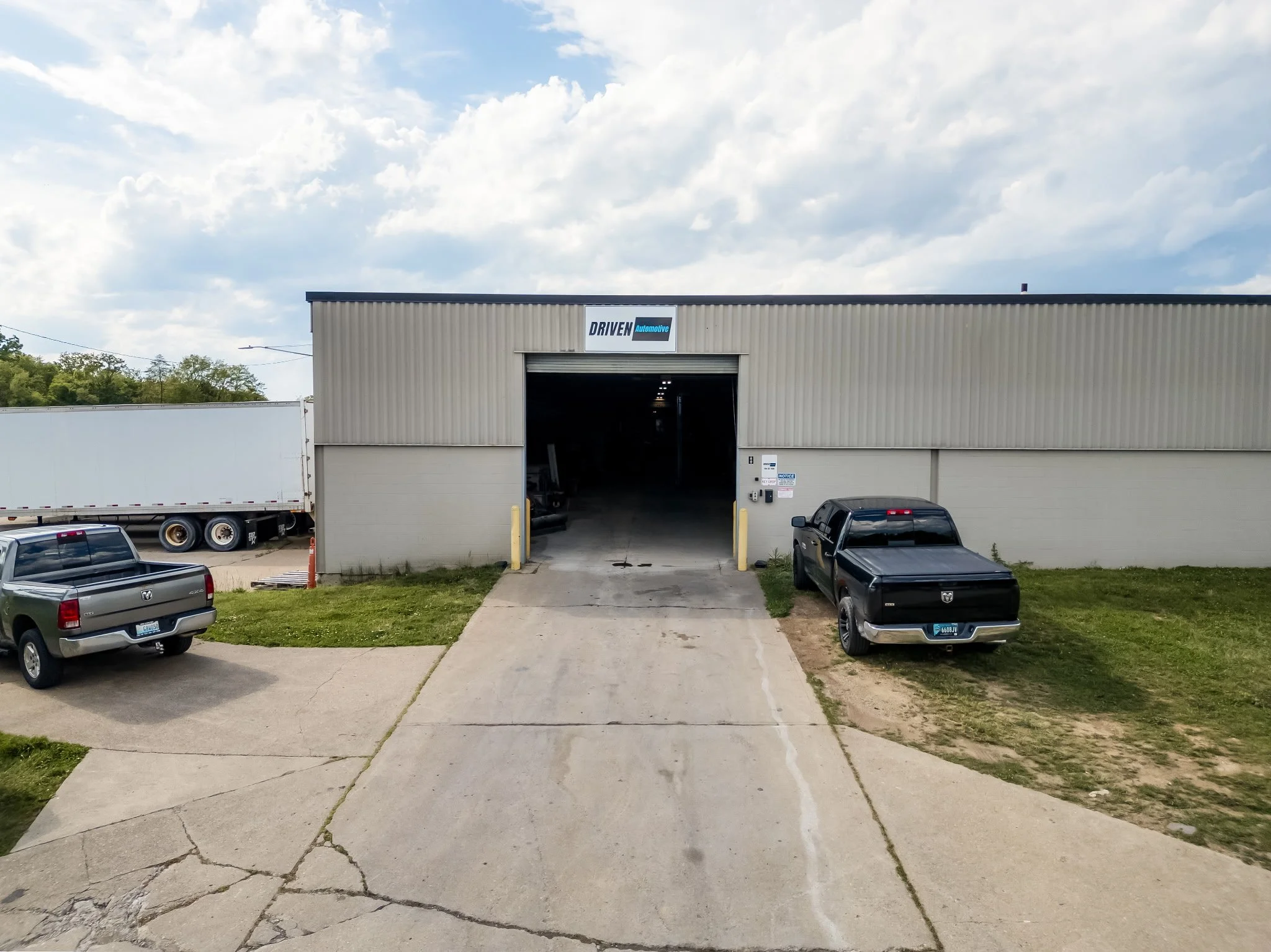A beige industrial building with an open garage door, a sign reading 'DRIVEN Automotive' on top, and parked trucks and cars in front. A cracked driveway leads up to the building with some grass on the sides under a partly cloudy sky.