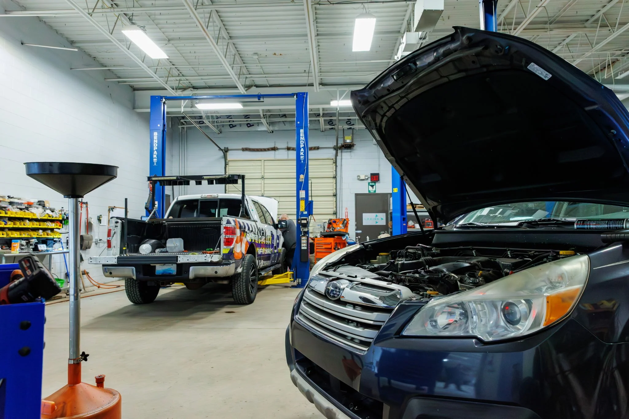 Auto repair shop with open hood of a gray car, another car on a lift, shelves with tools, and equipment.