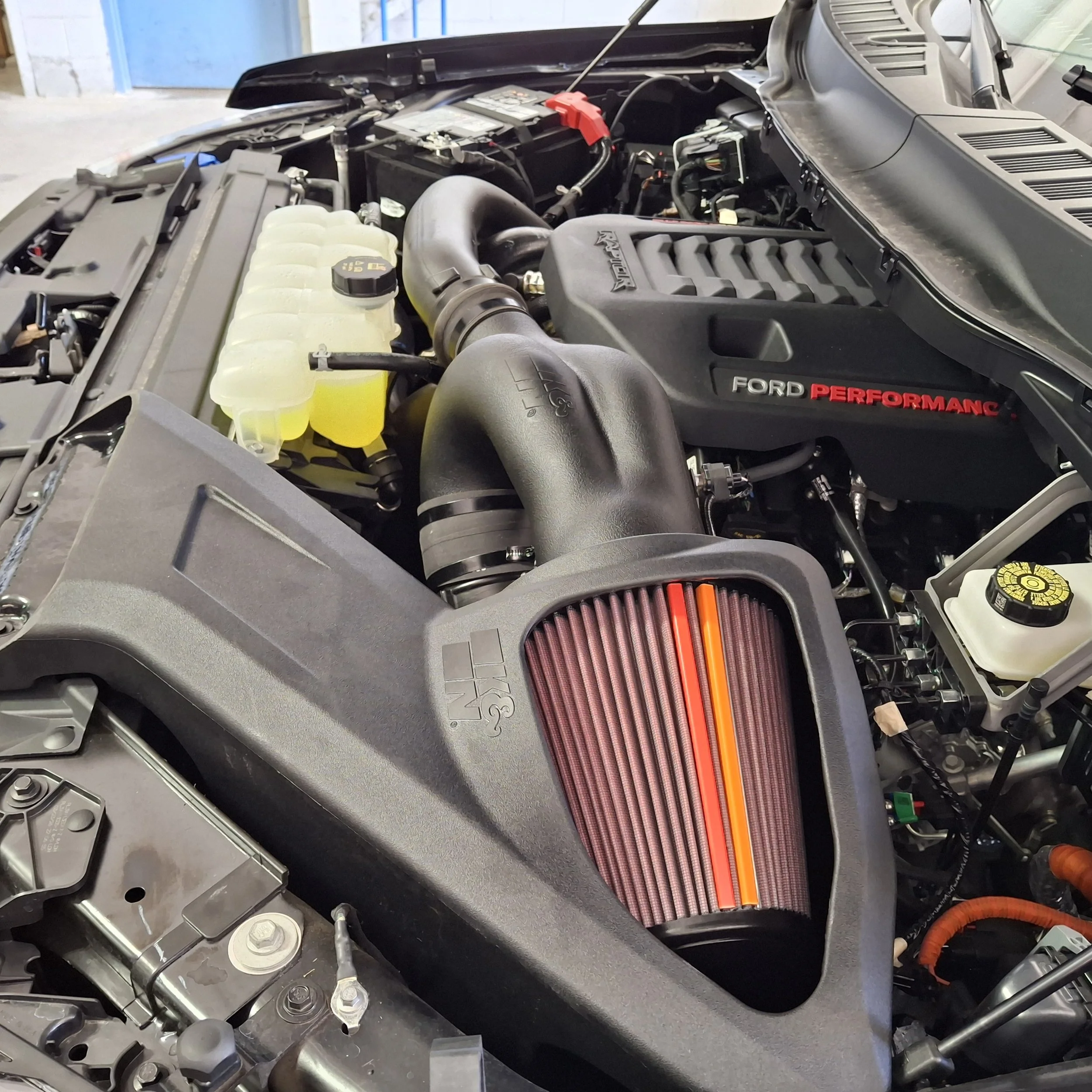 Close-up of a car engine bay with various components, including a red-topped performance air filter, radiator, and battery, in a garage.