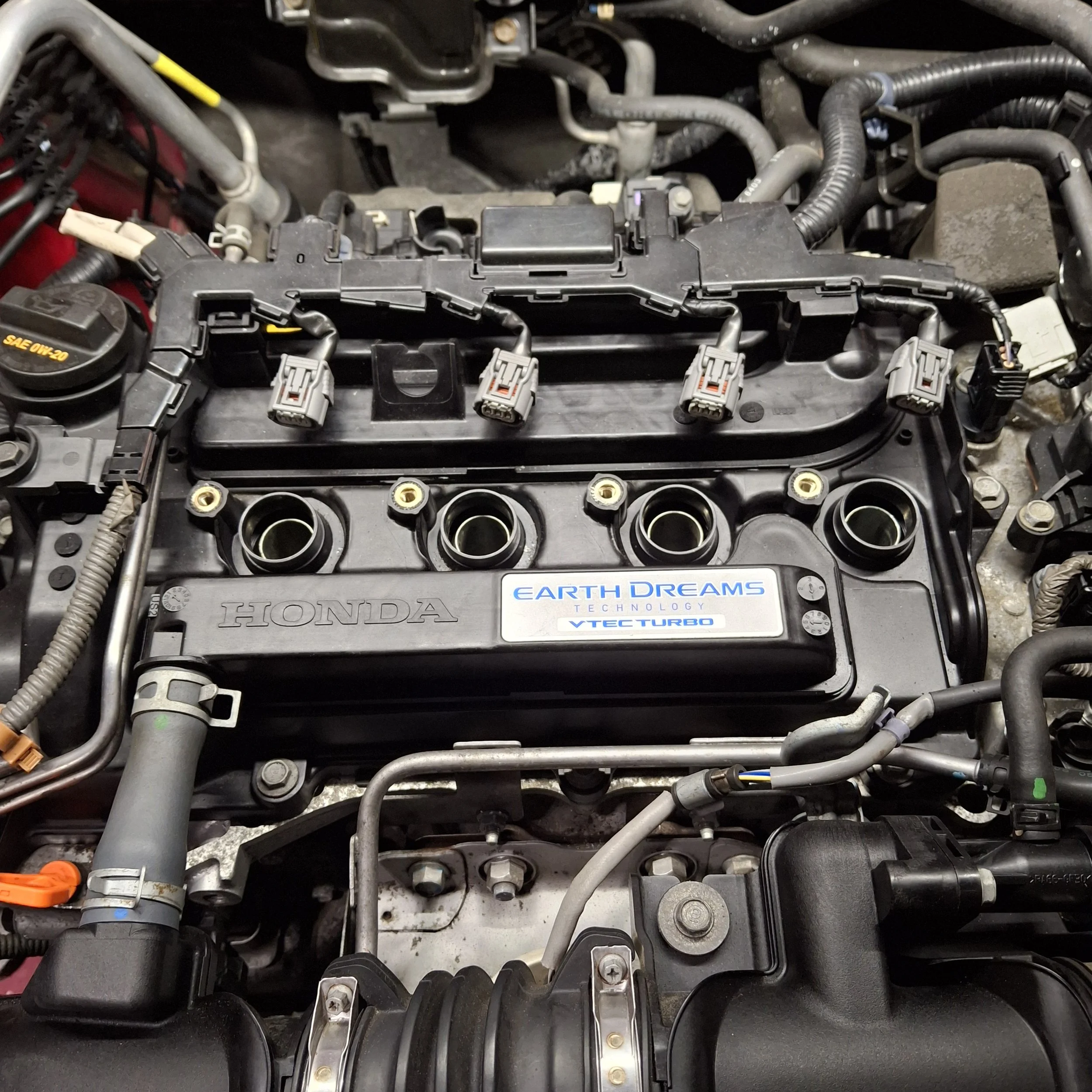 Car engine with various hoses, tubes, and electrical connectors visible, including a label that reads 'Earth Dreams Technology VTEC Turbo'.