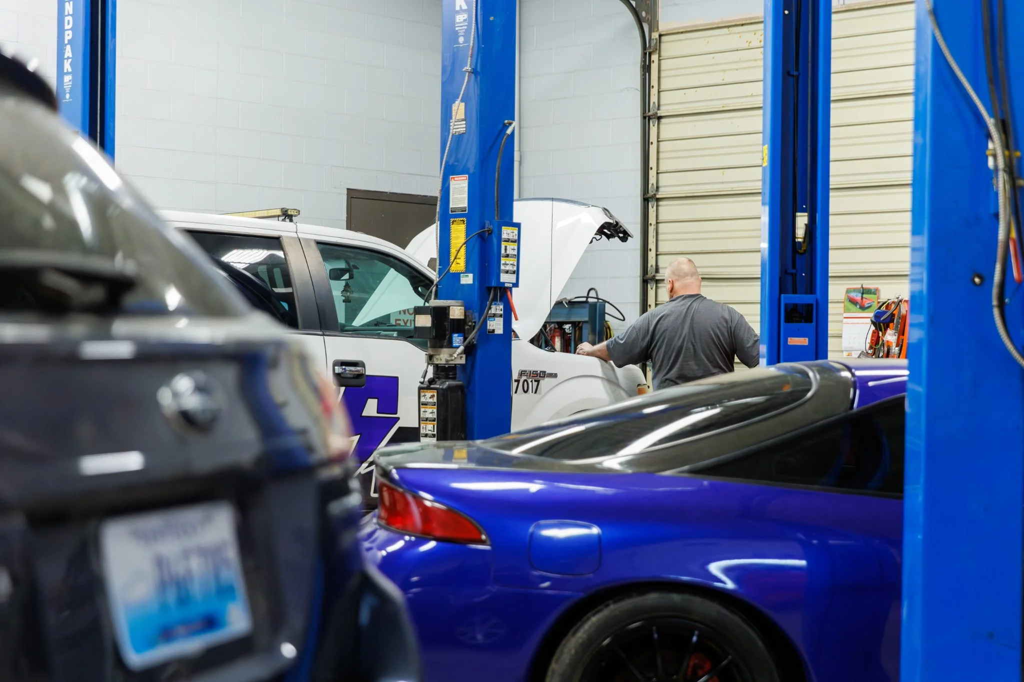 A mechanic working on a white vehicle inside an auto repair shop, with blue hydraulic lifts and various tools visible.