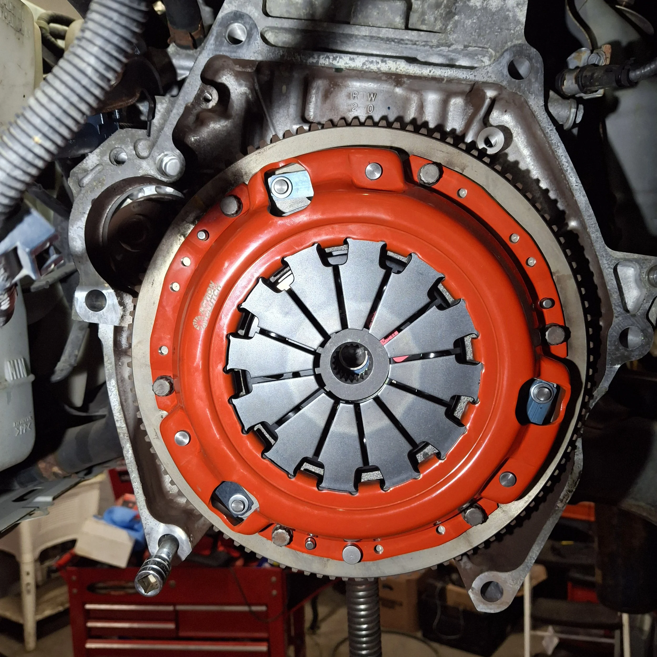 Close-up of a mechanical assembly featuring a large circular metallic component with a red outer ring, internal gear-like teeth, and black spokes. Various bolts and metal parts are attached, with some wires or tubes nearby, indicating it is part of machinery or automotive component.