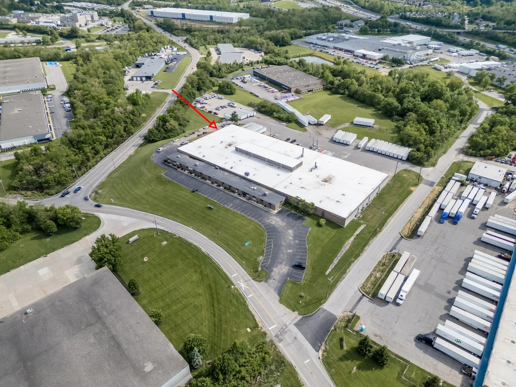 An aerial view of an industrial or commercial park with multiple buildings, parking lots, and green spaces, intersected by curving roads and surrounded by trees.