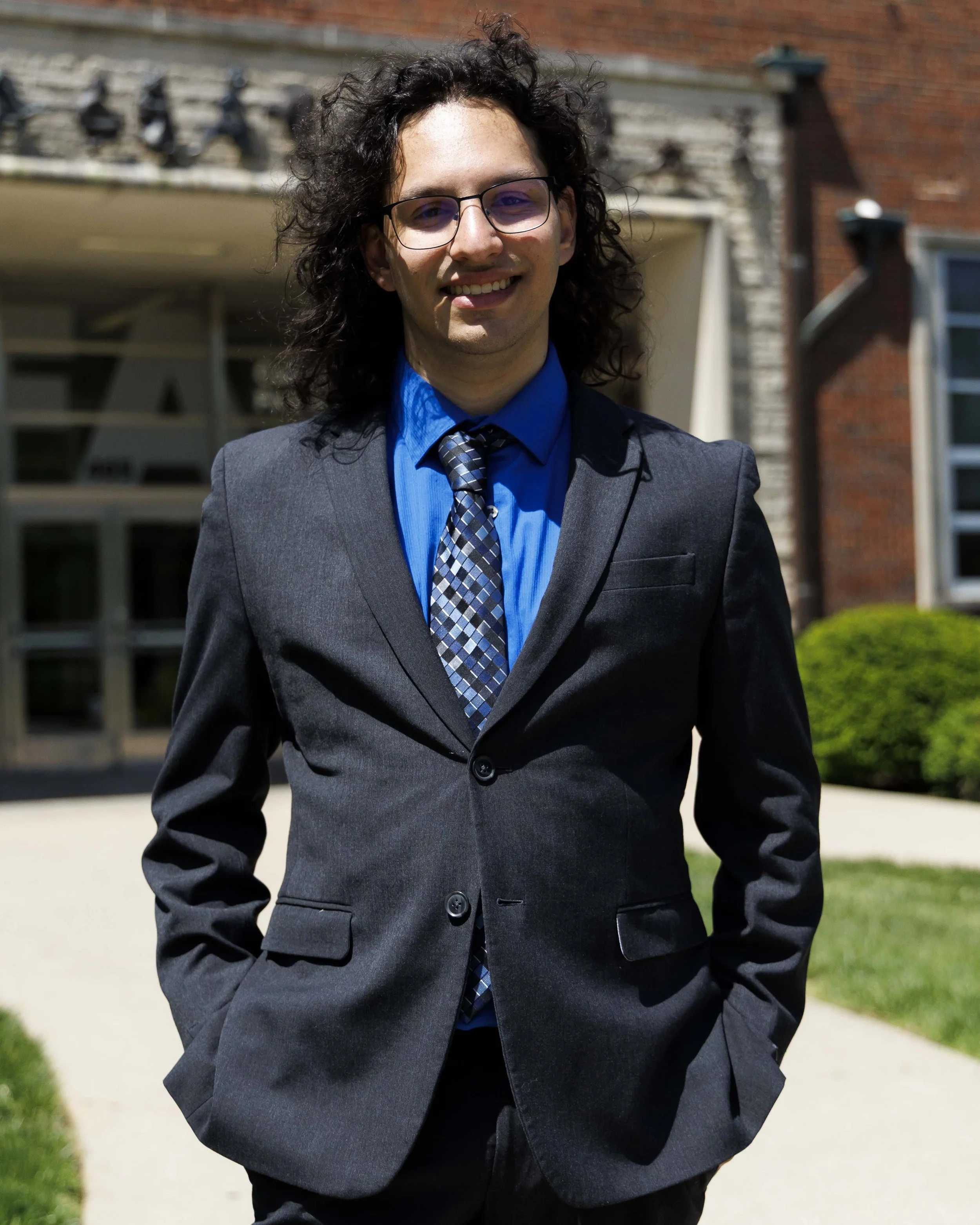 A smiling young man with curly dark hair and glasses wearing a dark gray suit, blue shirt, and patterned tie standing outdoors in front of a brick building with greenery.