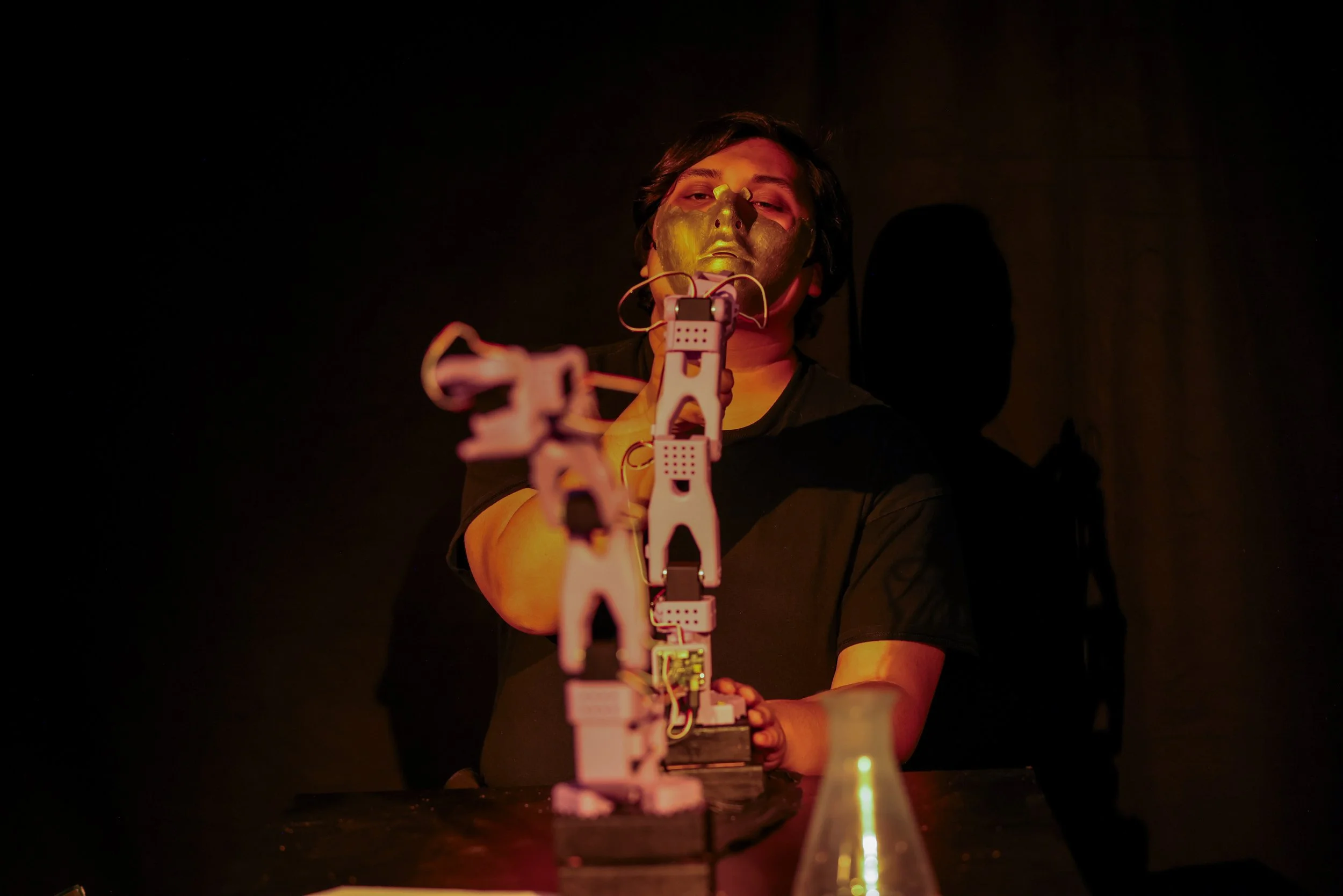 Person wearing a mask and sitting at a table with a firearm aimed forward, dim lighting with orange glow.