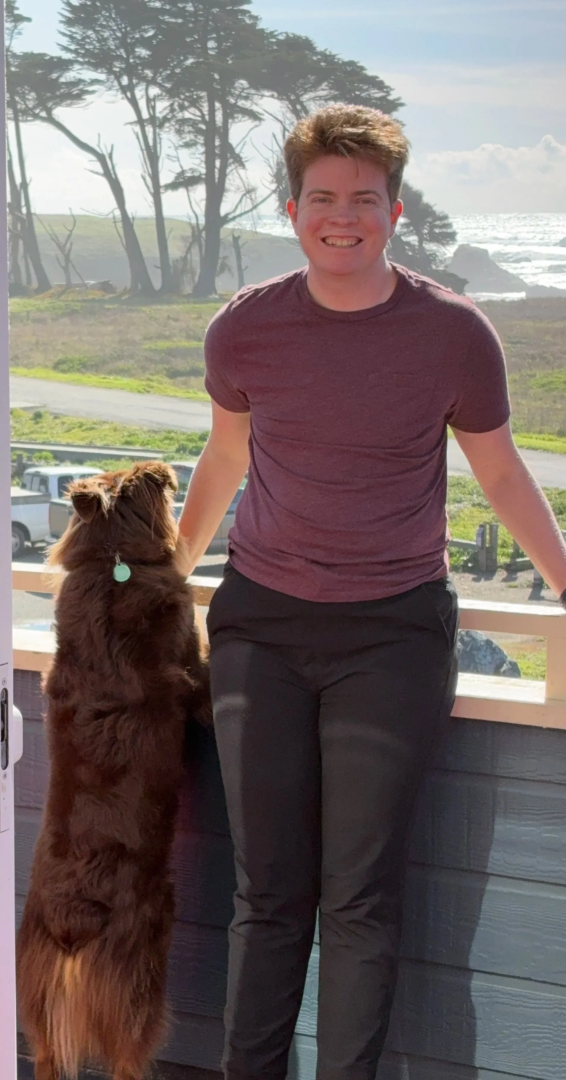 A non-binary human with light skin and short, light red hair standing outdoors next to a brown dog that is sitting on its hind legs and looking up at them. They are on a balcony with a scenic background of trees, grass, a coastal view, and the ocean.