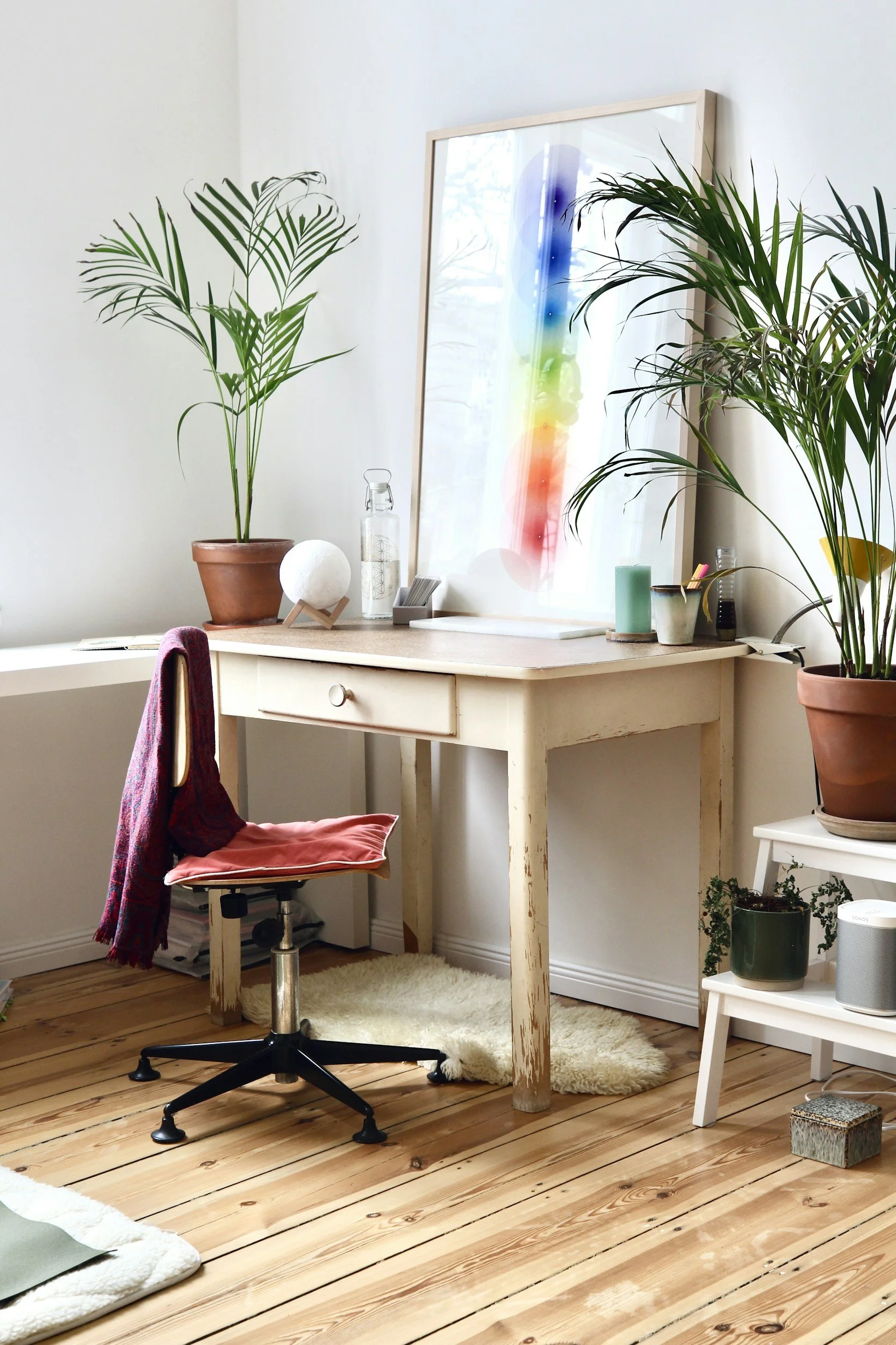 A home office workspace with a wooden desk, a swivel chair with a red scarf on it, a white shelf with potted plants, a large framed abstract rainbow art piece, and plants on either side of the desk against white walls and wooden floors.