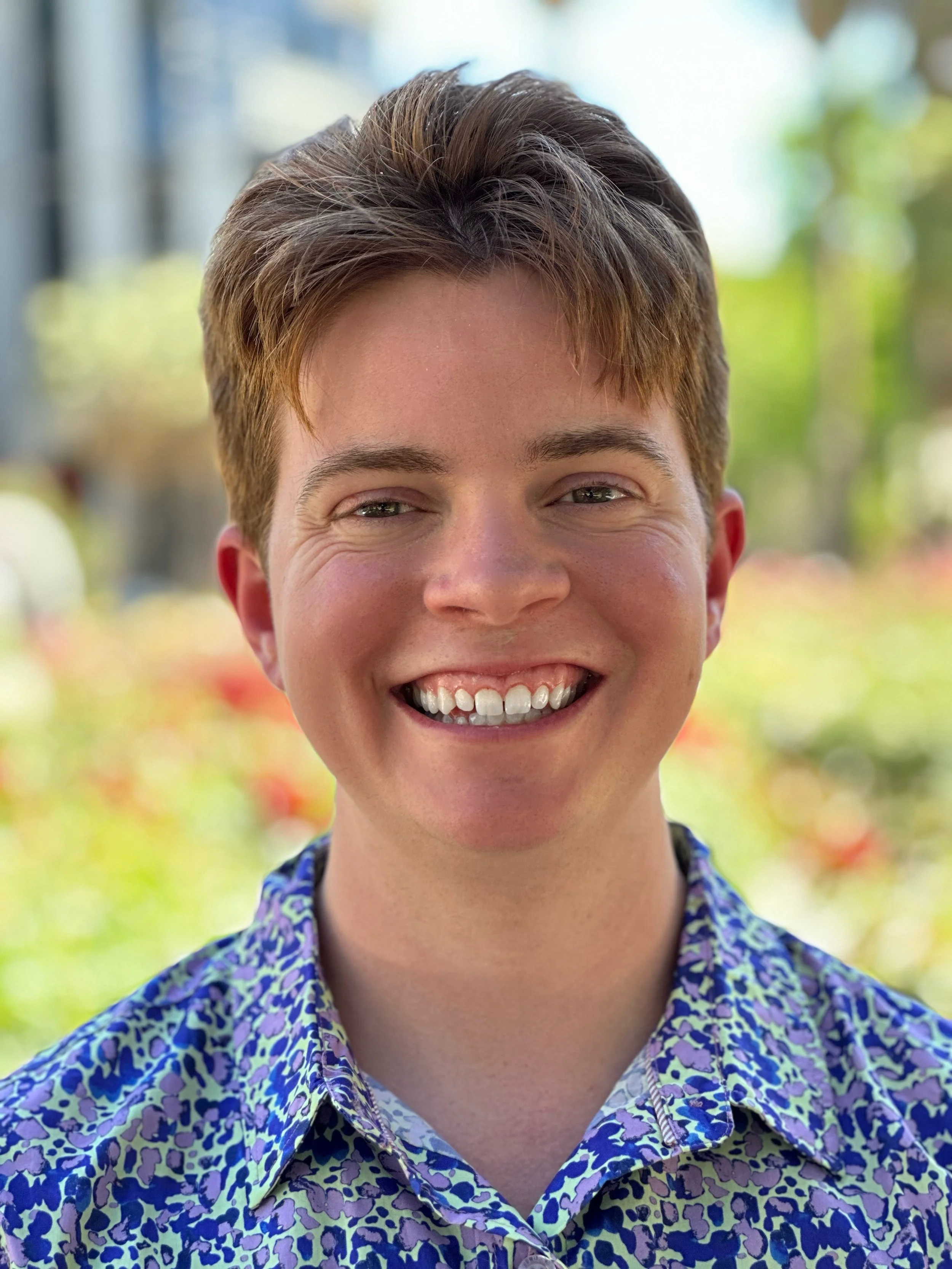 Close-up of a smiling young person with short red hair and a colorful patterned shirt, outdoors with blurred greenery and sunlight in the background.